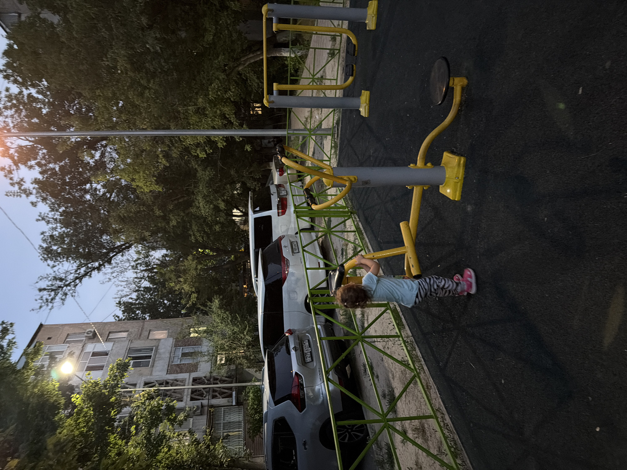 A young child, facing away from the camera, stands on an outdoor fitness equipment setup in an urban environment. The child is wearing a light blue t-shirt and striped leggings with pink shoes. They are holding onto the yellow handles of the equipment. The fitness equipment is yellow and grey, with a black disc at the end of one arm. Behind the equipment is a green metal fence. Beyond the fence, several cars are parked, including a silver SUV and two white minivans. In the background, a multi-story apartment building is visible, partially obscured by trees. The sky is a dusky blue, suggesting it is either early morning or late evening. The image appears to be taken in the city of Shymkent, Kazakhstan.