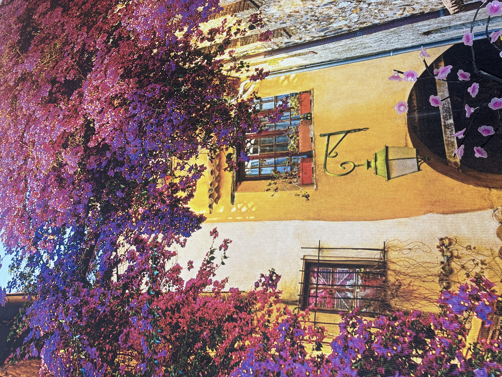 The image shows a vibrantly colored tapestry or artwork depicting a European-style building facade adorned with abundant purple and pink bougainvillea flowers. Two windows are visible: one on the left with wrought-iron bars and a window on the right with a wooden frame, both featuring planters with greenery. A vintage-style lamp hangs from the wall. The architecture suggests an old-world charm, possibly a Mediterranean or southern European setting. The scene is not located in Georgiyevsk, Russia, as there are no identifiable elements to support this claim.