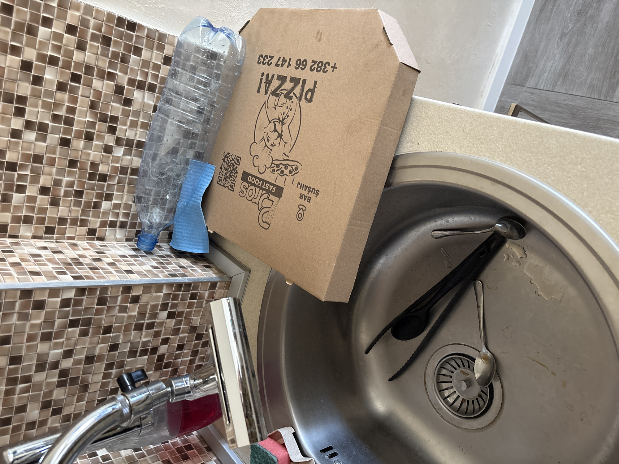 The image shows a cluttered kitchen sink area. A used plastic water bottle and blue cleaning cloth sit on a tiled surface next to a pizza box from "Bar Susanj, Bros Fast Food" (phone number +382 66 147 233). The sink contains a spoon, two metal spoons, and a black plastic cooking utensil. A faucet with a red soap dispenser attached is visible near the sink. The scene is likely in an apartment or home in Bar, Montenegro.