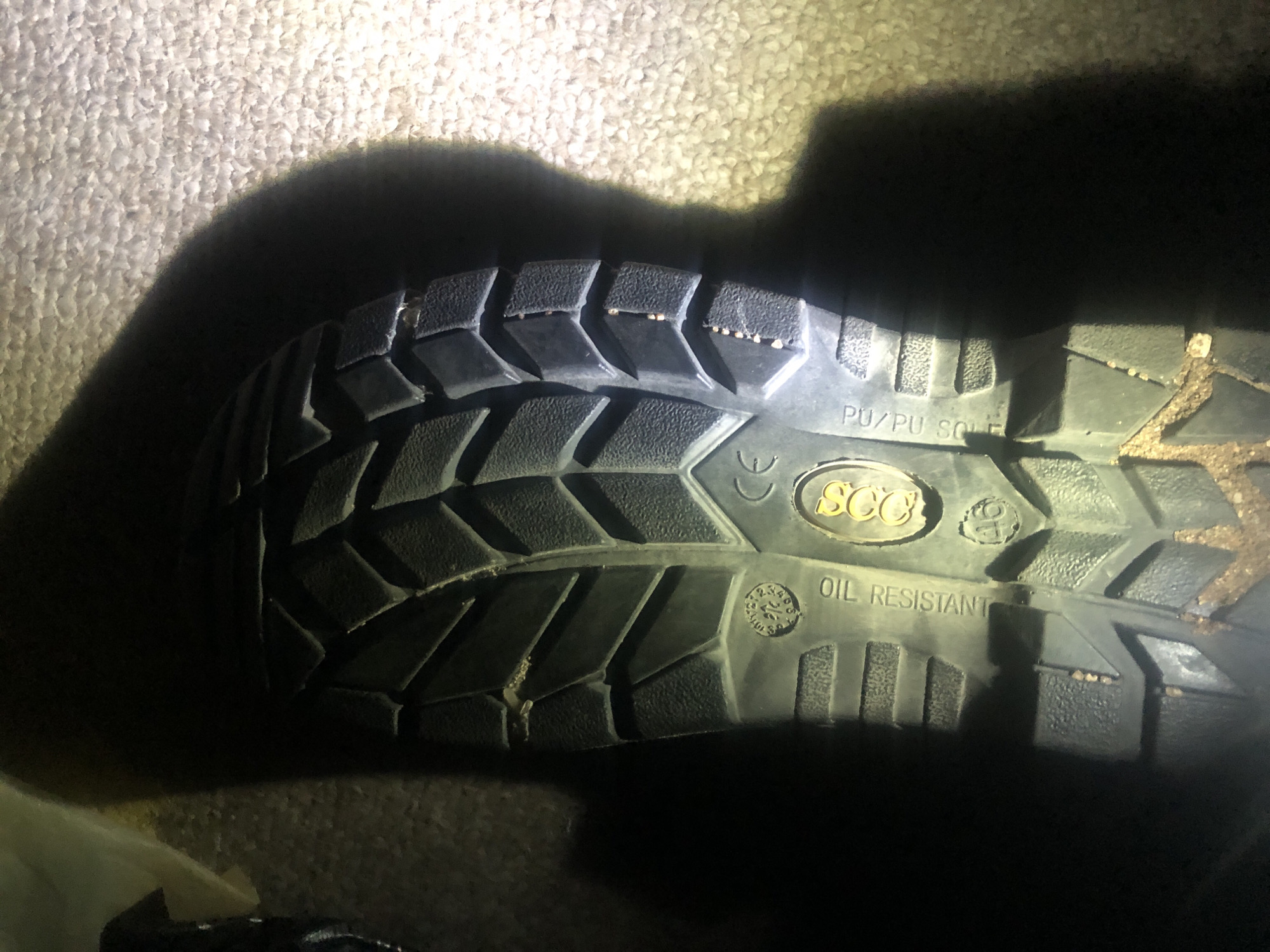 The image shows the sole of a black work boot resting on a carpeted surface. The boot sole has a deep tread pattern and several markings, including "OIL RESISTANT", "CE", and "PU/PU SOLE". There is a circular logo with the letters "SCO" inside it. The lighting in the image creates a strong shadow of the boot on the carpet. The scene is likely indoors, possibly in a residential or commercial setting. There is no indication of the location being Jalingo, Nigeria.