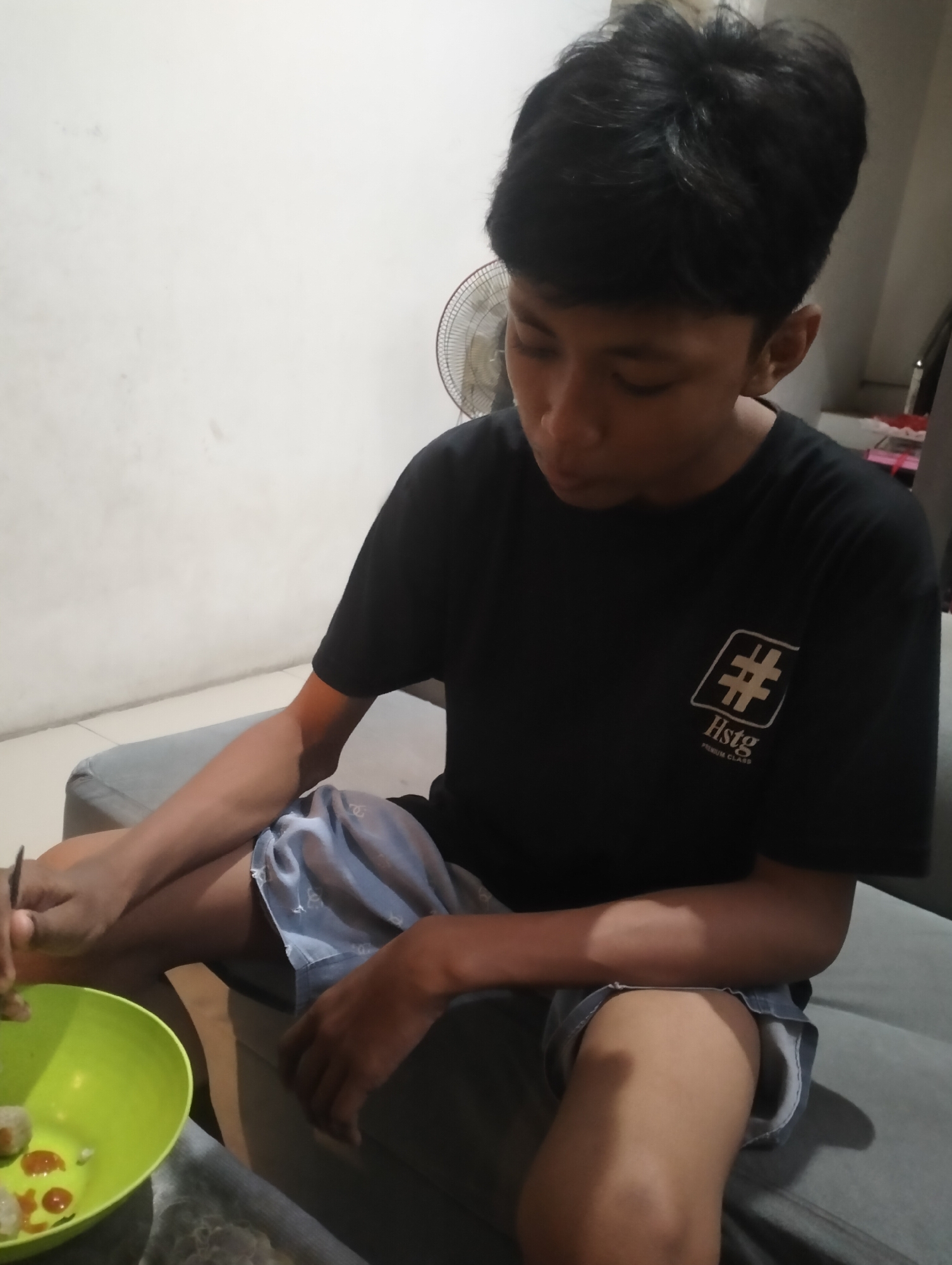 The image shows a young person sitting on a gray couch, looking down at a bright green bowl of food. The person is wearing a black t-shirt with a white logo that appears to be a hashtag symbol within a square, and the text "Hstg PREMIUM CLASS" below it. They are also wearing light blue shorts with a subtle pattern.
The person is holding a utensil, possibly a fork, in their left hand, and appears to be eating from the bowl, which contains several small, round food items with a reddish sauce. The lighting in the scene is dim, suggesting it might be evening or indoors with artificial light.
Behind the person, a white wall is visible, and a part of a round, caged fan can be seen on the left side of the frame. The overall setting appears to be a casual indoor environment, possibly a living room. The young person's expression is focused on the food, with no strong emotions immediately discernible. The location context provided is Sidodadi, Indonesia.