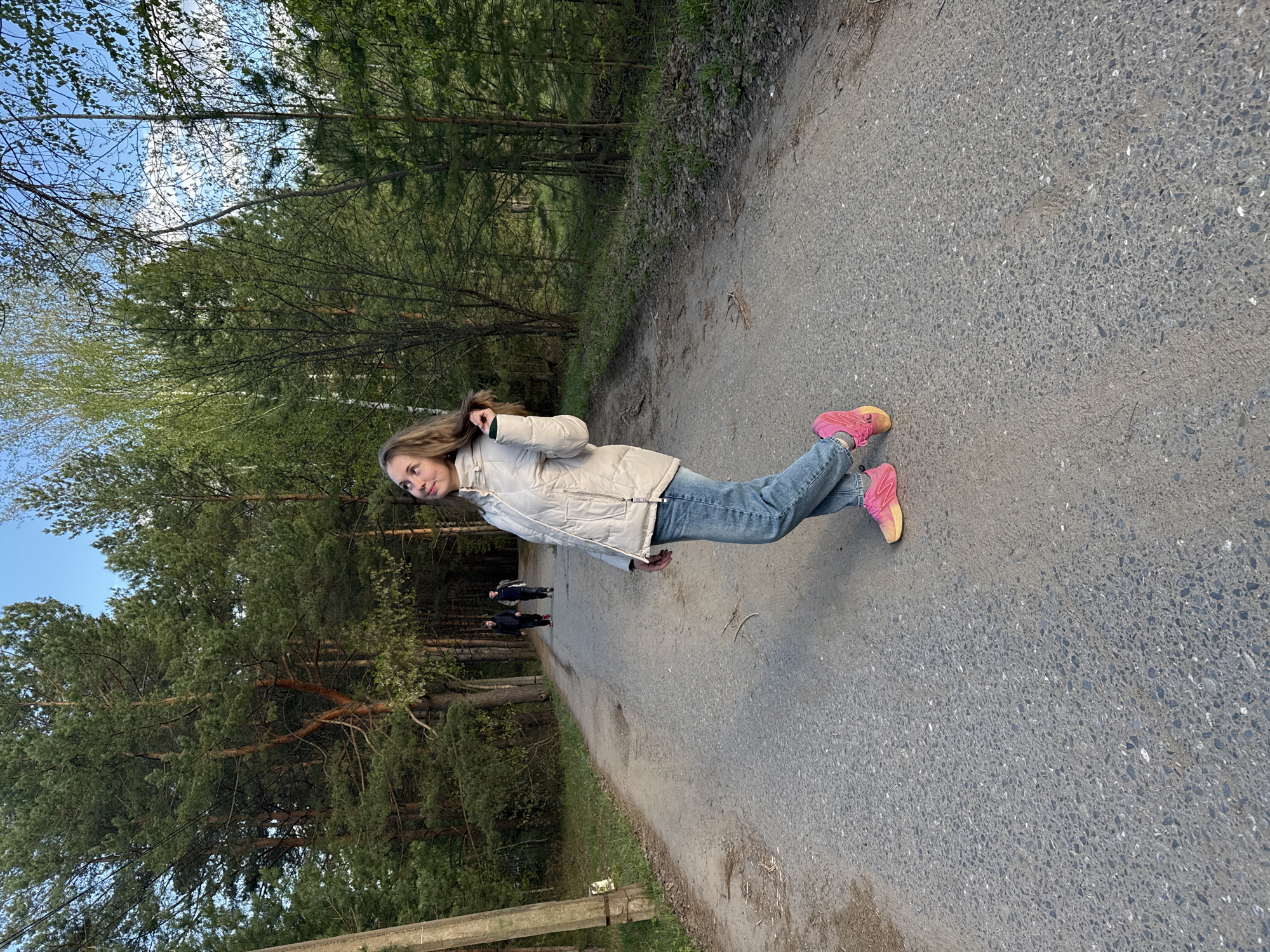 A woman with long brown hair is walking along a paved path in a park. She is wearing a white jacket, blue jeans, and bright pink sneakers. There are trees on either side of the path, and two people can be seen walking behind her in the distance. The path appears to be in the city of Unknown, Unknown.