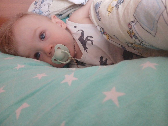 A young child, appearing to be an infant or toddler with fair skin, light hair, and blue eyes, is depicted lying on their left side in an indoor domestic setting. The child is looking directly towards the viewer and has a light green pacifier in their mouth, from which a thin, dark chain or cord extends. The child wears a white top or onesie patterned with dark silhouettes, possibly of dinosaurs. They are resting on a light blue-green sheet or blanket adorned with multiple white star patterns. A separate white blanket, decorated with colorful cartoon characters (resembling Winnie the Pooh and friends), is draped over a portion of the child's body. The specific location within the city of Chaikovsky, Russia, is not discernible from the visual information provided.