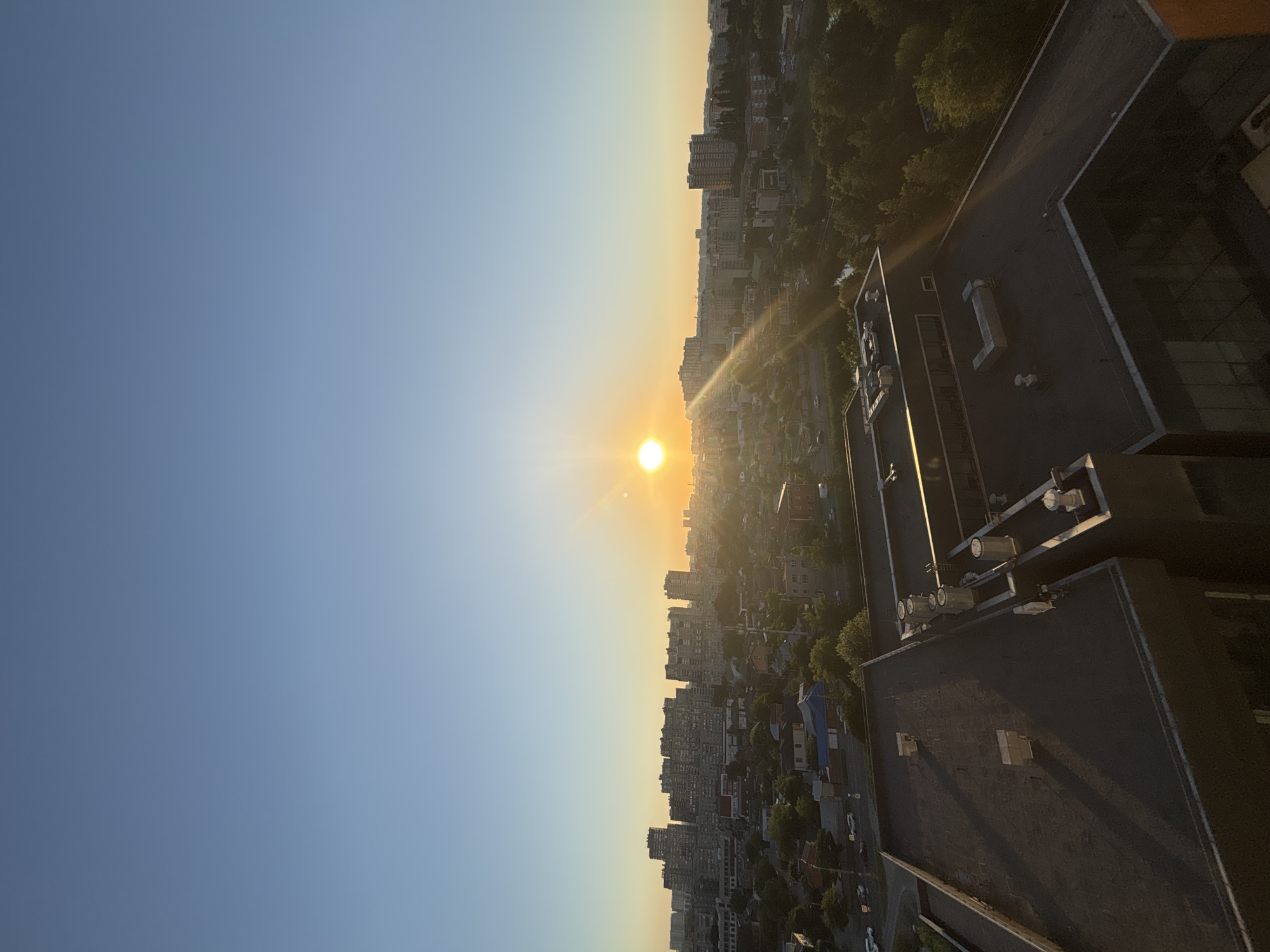 The image depicts a panoramic view of a city skyline at sunset, seen from a high vantage point, likely a rooftop. The sun, a bright, warm orb, is positioned on the left side of the frame, casting long shadows and creating a warm, golden glow across the sky and cityscape. The sky transitions from a clear, pale blue on the left to a soft, hazy yellow-orange near the horizon.
The city is a dense collection of buildings, with a mix of high-rise apartment blocks and smaller residential structures. Trees are interspersed throughout the urban landscape, adding a touch of green to the predominantly grey and beige tones of the buildings. In the foreground, the dark, flat roofs of several industrial or commercial buildings are visible, complete with ventilation units and other rooftop equipment. One of these buildings has a stark white outline along its edge.
The scene appears to be peaceful and serene, capturing the quiet beauty of a city transitioning from day to night. The time of day is clearly sunset, indicated by the low angle of the sun and the warm, diffused light. The weather appears clear and fair, with no visible clouds. The location is suggested to be Krasnodar, Russia, based on the provided context. There is no visible text in the image. The overall impression is one of urban sprawl bathed in the atmospheric light of dusk.