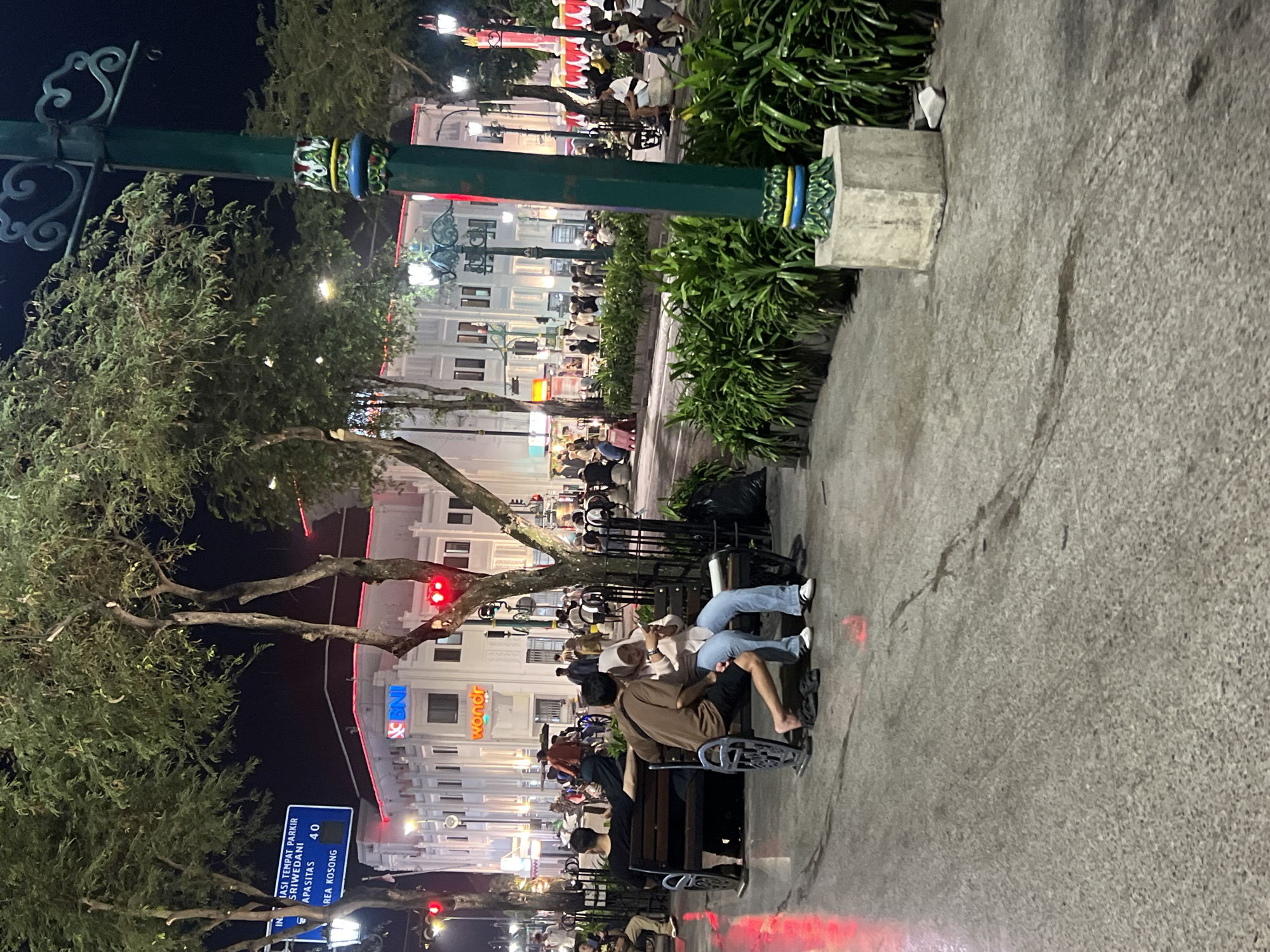 The image depicts a nighttime scene at Malioboro Street in Yogyakarta, Indonesia. A paved pedestrian walkway is visible, with a dark gray, speckled surface showing wet patches, likely from recent rain or street cleaning. Several people are present, engaged in various activities. In the foreground, a couple is seated on a dark metal bench. The man is wearing a brown shirt and dark shorts, with his left leg bare and bare feet resting on the ground. The woman is wearing a light pink long-sleeved shirt and light blue jeans, with her head covered by a beige hijab. They appear to be interacting with a phone. To their left, another person is seated on the same bench, wearing a black shirt.
Behind the bench, a row of lush green bushes forms a boundary. A tall, dark green lamppost with decorative elements on its base stands prominently in the right mid-ground. In the background, a multi-story white building with red neon lights outlining its upper floors is visible, with signs that read "BNI" and "wondr." The building is illuminated, and several streetlights are also on, casting a glow on the scene. Numerous other people are scattered throughout the background, some walking and others gathered in groups. A blue street sign with white text is visible in the upper left, indicating directions. The overall atmosphere is one of a busy urban street at night, with a mix of local residents and possibly tourists.