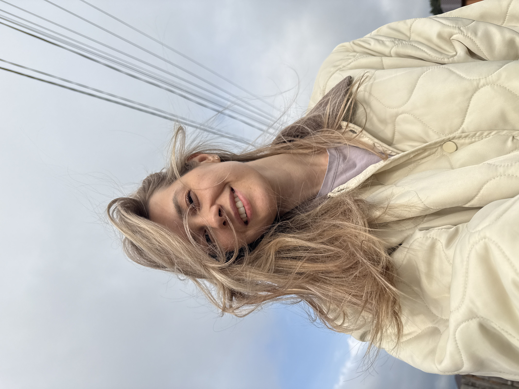 A woman with long blonde hair is smiling at the camera while wearing a light-colored, quilted jacket. The background includes an overcast sky and overhead power lines. The image was taken in the city of Chaikovsky, Russia.