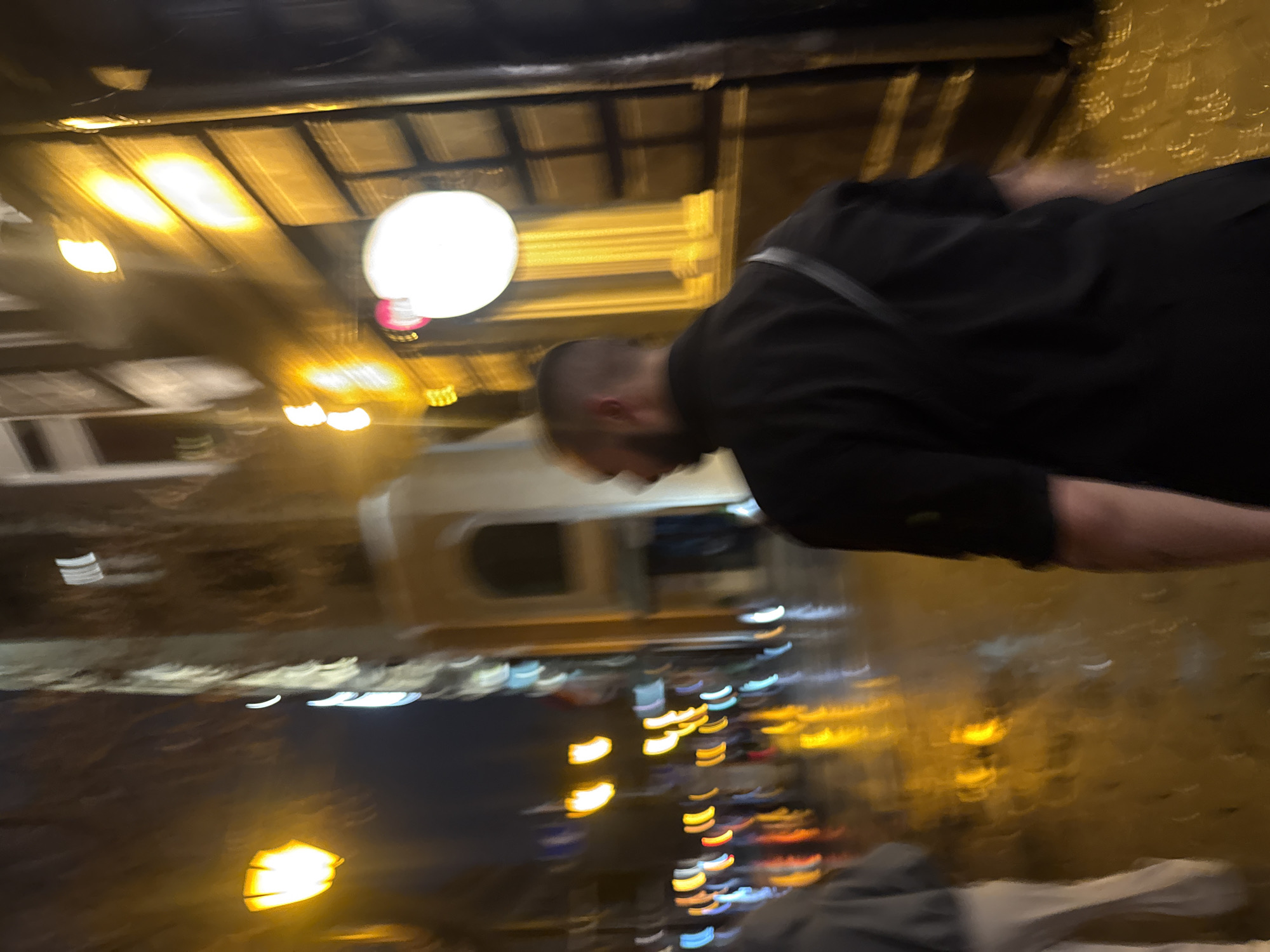 The image is a blurred, slightly off-kilter shot taken at night. A man with a shaved head wearing a black jacket is centrally positioned, though his face is obscured. The background suggests an urban setting, likely in Yerevan, Armenia, with a variety of light sources creating a bokeh effect. There are lights from buildings, streetlights, and possibly vehicle headlights. The general ambience is dark and potentially wet, given the light reflections. A ceiling with visible lighting fixtures is present at the top of the frame.
