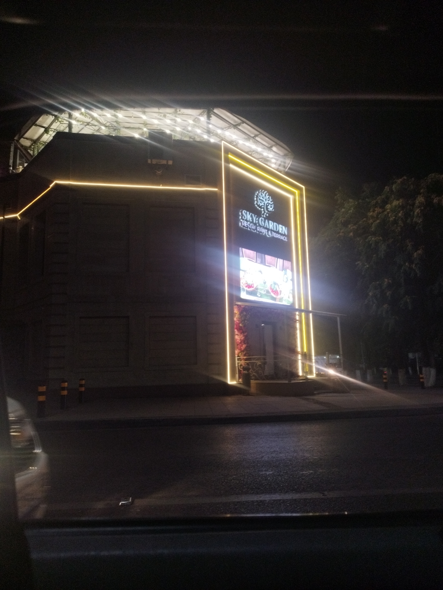 The image shows the "Sky Garden Restaurant & Terrace" located in the city of Shymkent, Kazakhstan. The building is illuminated at night with bright yellow neon lights outlining its facade and a large digital display screen. The screen shows vibrant imagery, possibly advertisements or decorative visuals. The surrounding environment appears to be a street with a sidewalk and some trees visible in the background. The overall impression is of a modern establishment with eye-catching night-time lighting.