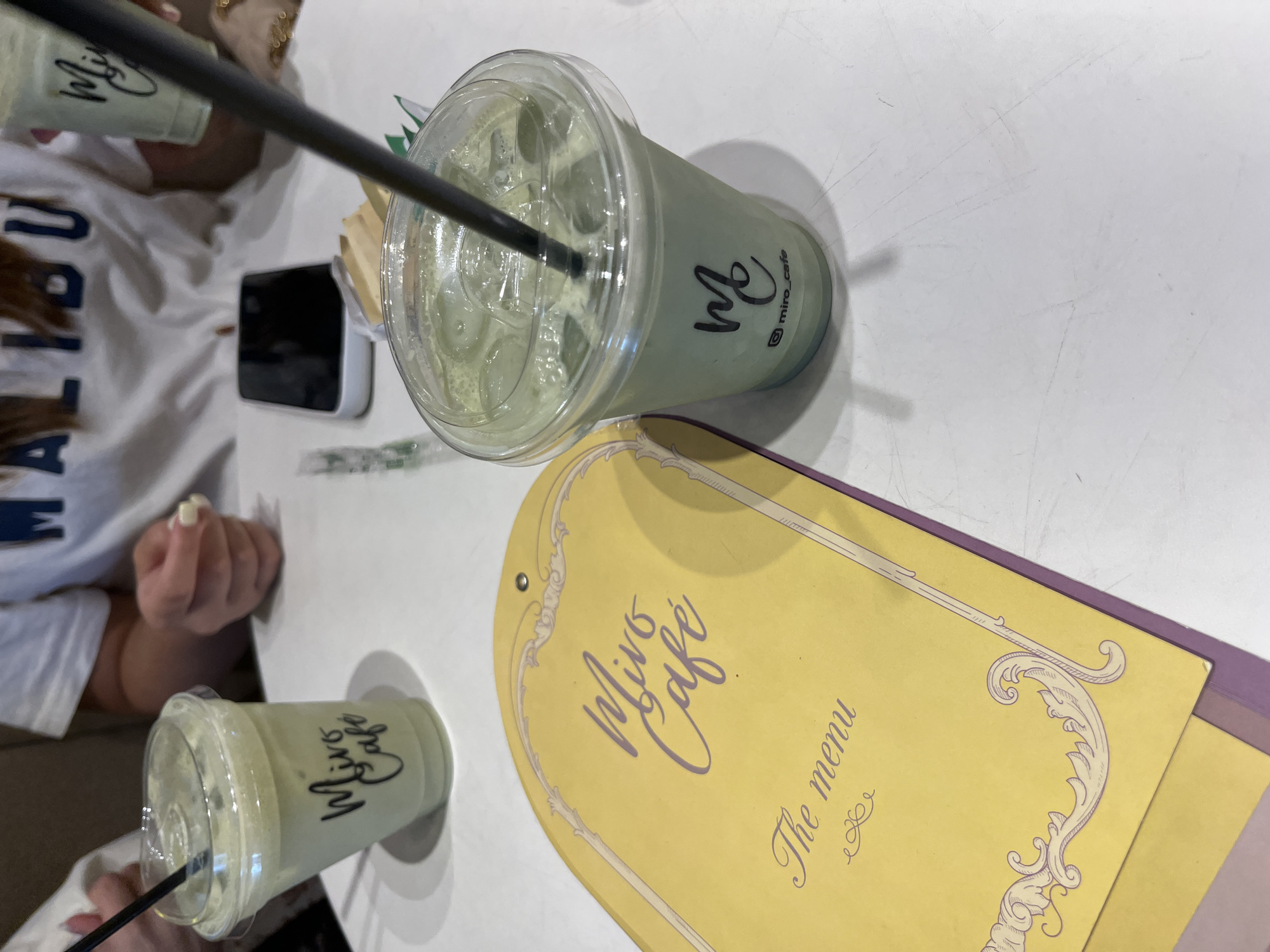 The image depicts a table at Miro Cafe in Puebla, Mexico. Two iced matcha lattes, in clear plastic cups with black straws, sit on the white table. A yellow Miro Cafe menu with purple trim is partially visible. An iPhone is also on the table. A person wearing a white t-shirt with "MALIBU" printed on it is partially visible to the left, with a hand near the drinks.
