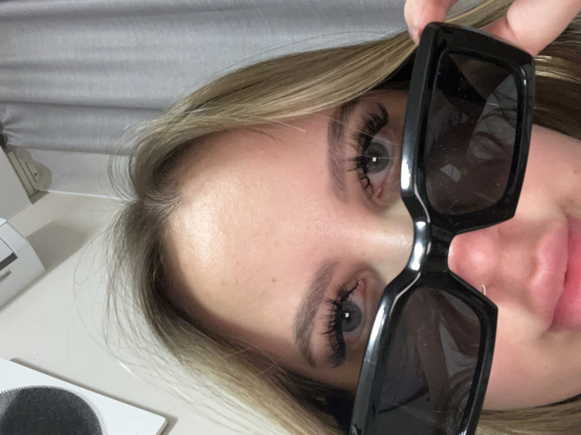 A close-up, rotated image shows a woman with blonde hair, blue eyes, and false eyelashes holding a pair of large, black sunglasses in front of her face. The background is a light grey curtain. To the left, a white surface with a partially visible circular black object is present. There is no visible evidence indicating the location is Krasnodar, Russia.