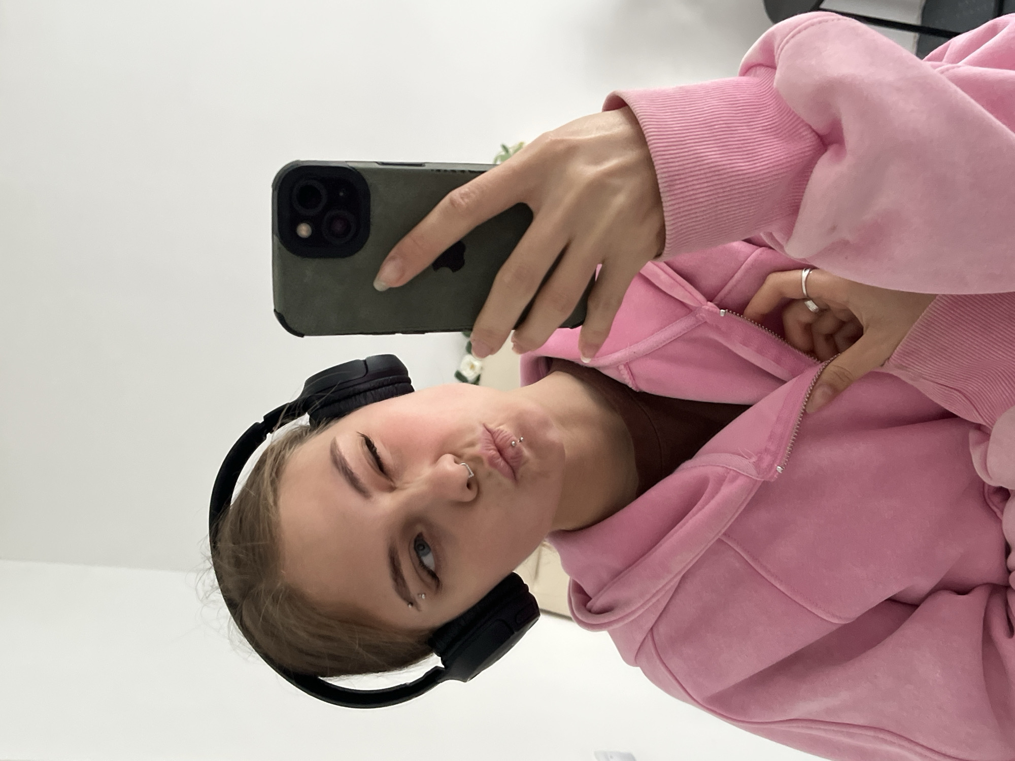 A woman is taking a selfie indoors. She is wearing black headphones and a pink hoodie. She has piercings in her nose, lip, and eyebrow. She is holding a smartphone with an olive-green case. The location is unknown, as the prompt incorrectly mentions a city in Russia unrelated to the image.