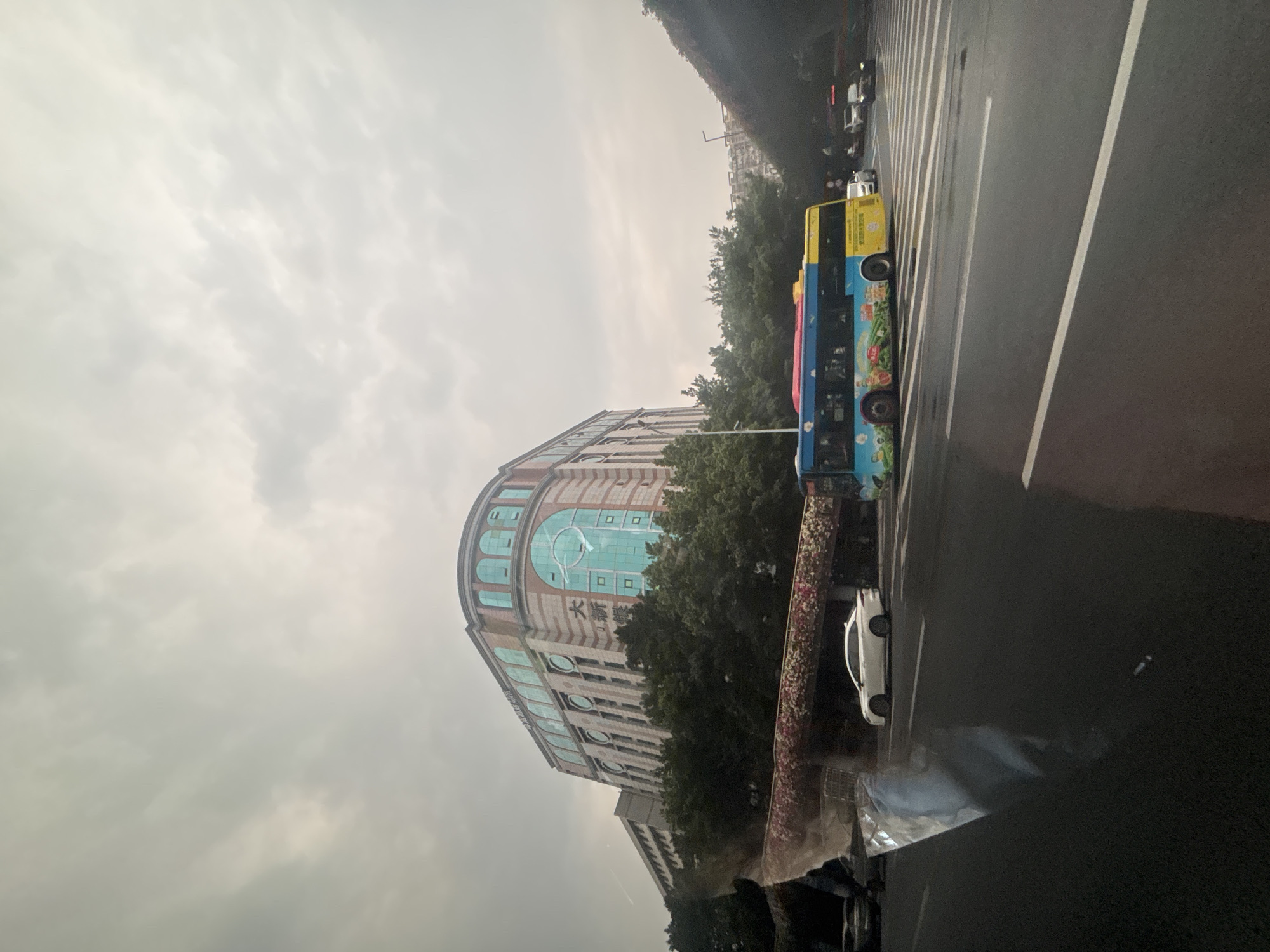 The image captures a street scene in Yuexiu District, China, under an overcast sky. In the background, a large, multi-story building with arched windows and decorative bands of red and white stripes dominates the view. The building appears to have a semicircular or curved facade with several stories. In front of this building, a dense line of green trees forms a natural barrier.
A brightly colored bus, predominantly yellow and blue with colorful illustrations, is the central focus of the mid-ground. The bus is positioned on the right side of the street, in motion. To the left of the bus, a white sedan is also on the road. The street is a multi-lane roadway with visible white lane markings and a pedestrian crossing.
In the foreground, the dark asphalt of the road is visible, with reflections of the sky and possibly vehicles on its wet surface, suggesting recent rain. There are also some indistinct reflections and distortions in the lower left portion of the image, potentially from a window or a surface within the vehicle from which the photo was taken. Further elements of the urban environment, such as streetlights and other vehicles, are visible in the distance.