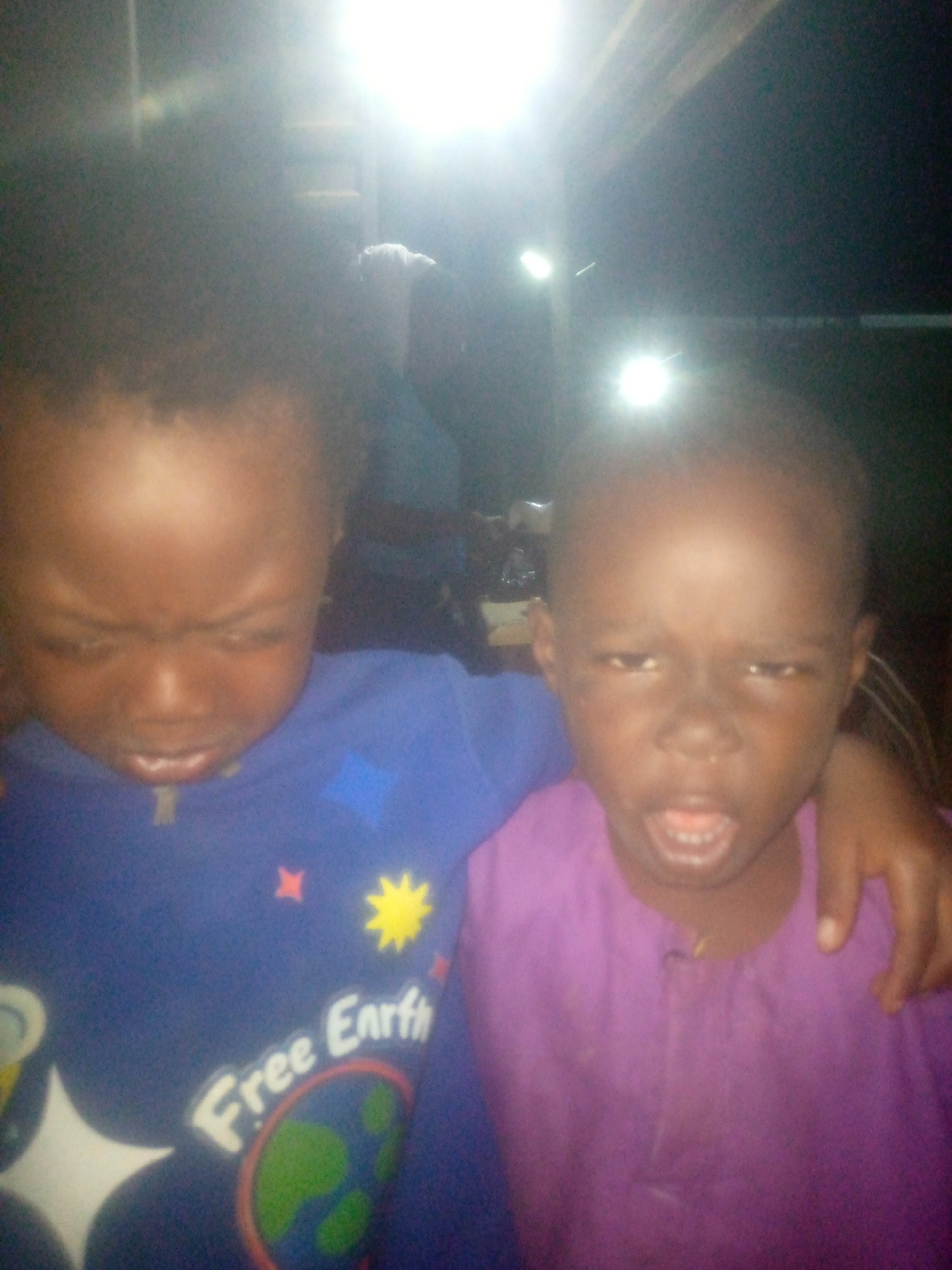 The image features two young children, likely boys, captured in a poorly lit environment, possibly indoors or at night, with prominent bright light sources in the background.
The child on the left wears a blue long-sleeved shirt. Their head is tilted down, and their eyes are squinted, suggesting a reaction to the bright light or a deliberate facial expression. The shirt has a graphic design including a yellow sun, a red star, and a depiction of the Earth with the white text "Free Earth" clearly visible below it.
The child on the right stands beside and slightly behind the first child, with an arm resting on the left child's shoulder, indicating closeness. This child wears a purple long-sleeved garment, possibly a traditional tunic, and has an open mouth and squinted eyes, also seemingly reacting to the intense light.
The background is dark and out of focus, with several bright, glaring light sources creating significant overexposure and lens flare. A blurry figure can be vaguely seen further back. The overall lighting conditions result in the subjects appearing underexposed and slightly blurry, making definitive identification of facial expressions challenging beyond the general squinting. The photo captures a candid moment between the two children.
