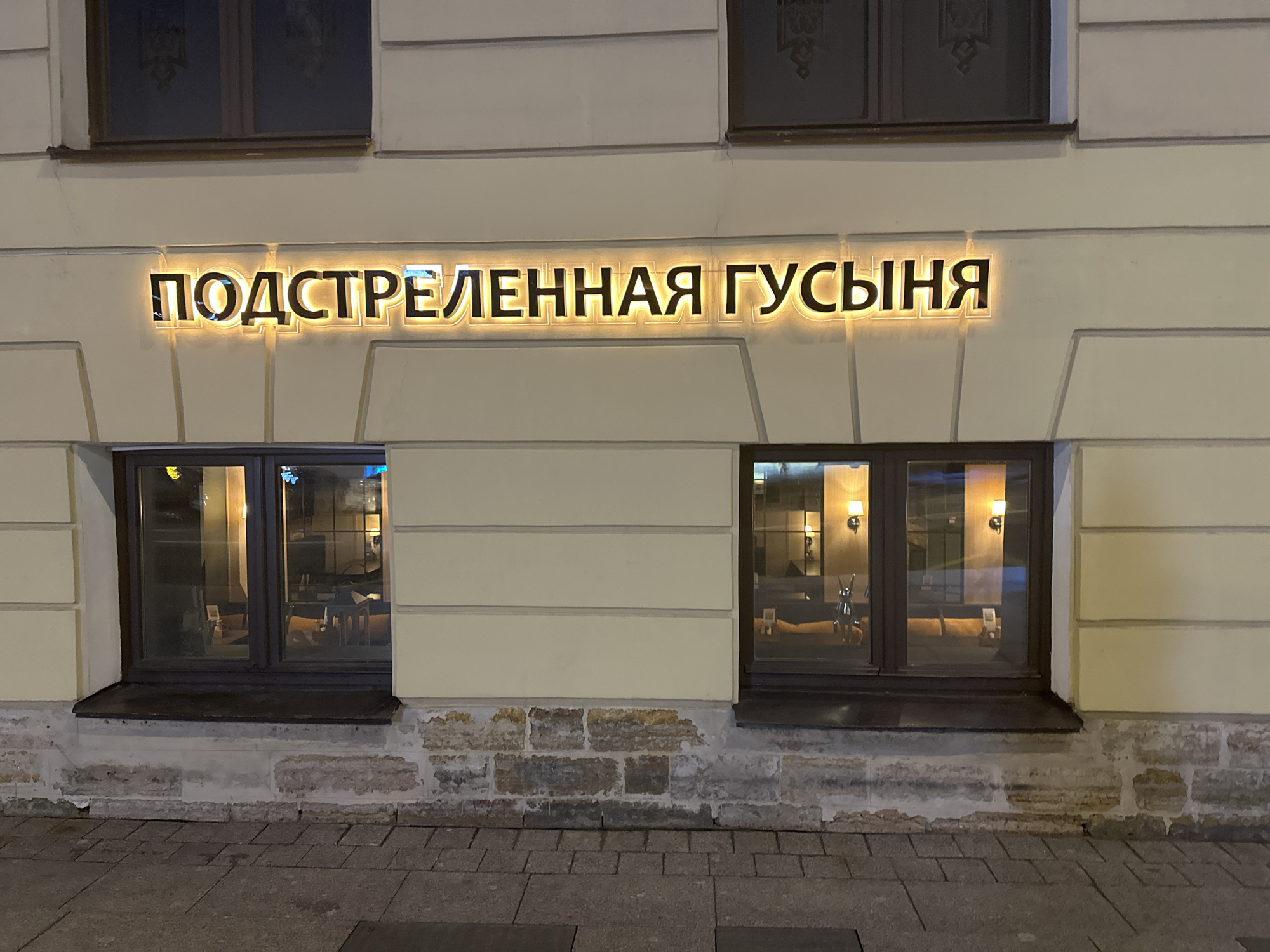 The image shows the facade of a building, with a lit sign reading "ПОДСТРЕЛЕННАЯ ГУСЫНЯ". There are two windows, and the lower part of the facade is made of stone. The image was taken in Saint Petersburg, Russia.