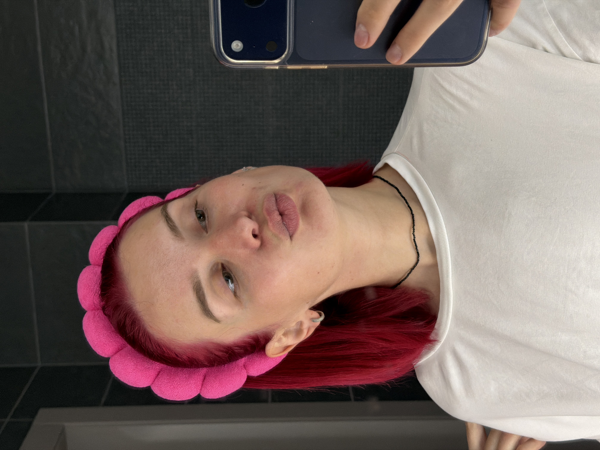 A person with red hair and a pink headband is holding a smartphone to take a selfie. The person is wearing a white t-shirt and a black beaded necklace. The background consists of dark gray, square tiled walls and a white bathroom sink. There is no indication in the image that the location is in Chaikovsky, Russia.