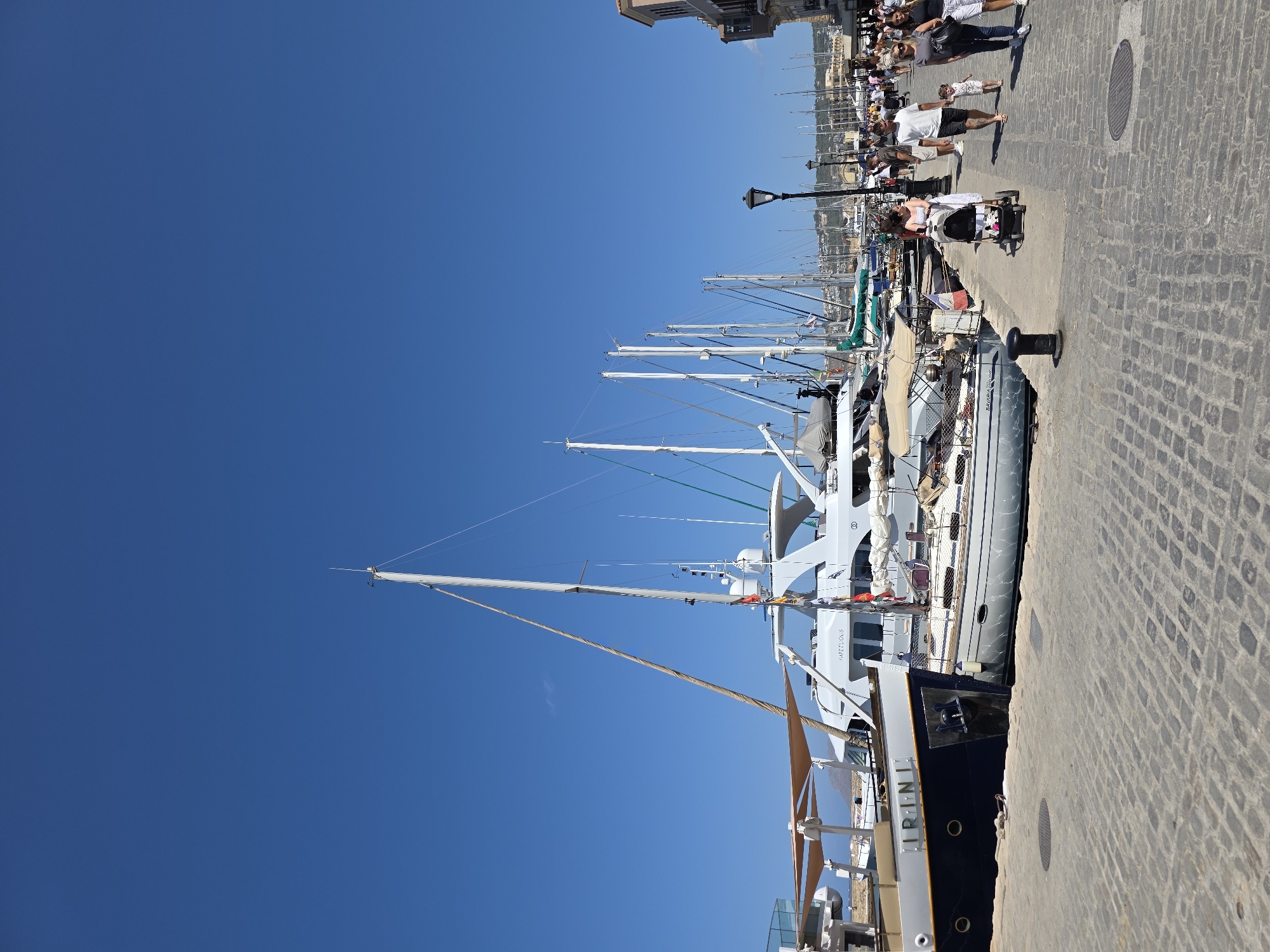 The image shows a harbor scene in Chania, Greece, on a clear, sunny day. Several sailboats and yachts are docked along a cobblestone promenade. In the foreground, a dark-hulled boat with the name "IRINI" is visible, alongside a smaller sailboat. Further down the promenade, a group of people are walking, including a woman pushing a stroller. Buildings with traditional architecture line the right side of the promenade, suggesting a historic urban setting. The sky is a vibrant, cloudless blue.