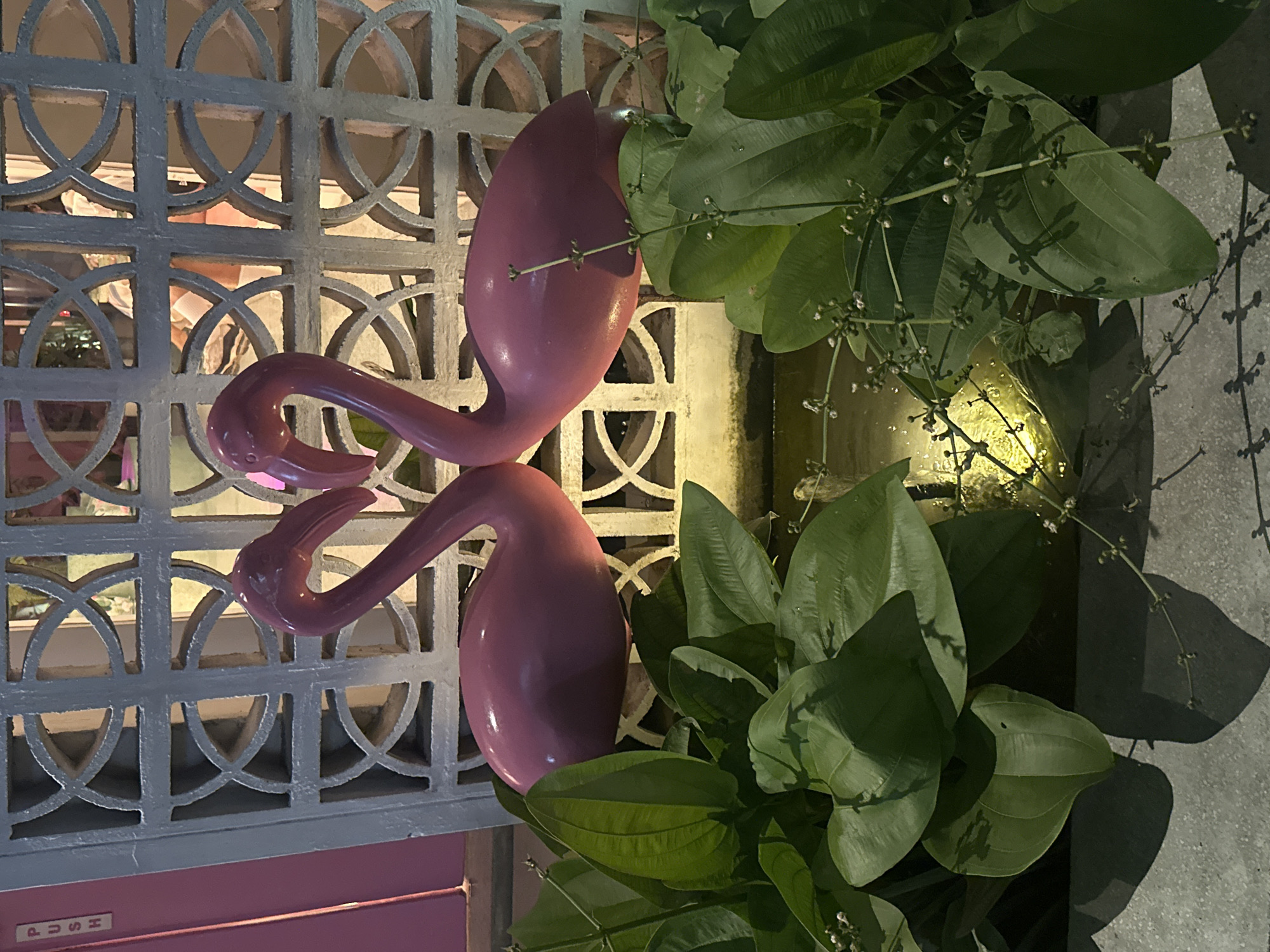 The image depicts a close-up shot of a decorative scene, likely located in North Kuta, Indonesia. Two pink flamingo sculptures are arranged vertically, one above the other, with their heads facing in opposite directions. They are positioned against a decorative gray wall with a repeating geometric pattern of interlocking circles and squares. To the right of the flamingos are large, green, heart-shaped leaves of a plant, possibly growing in a nearby planter. The lighting suggests an evening or nighttime setting, with some areas cast in shadow and others illuminated. The overall composition is colorful and aesthetically pleasing, suggesting a decorative element in a restaurant or shop.
