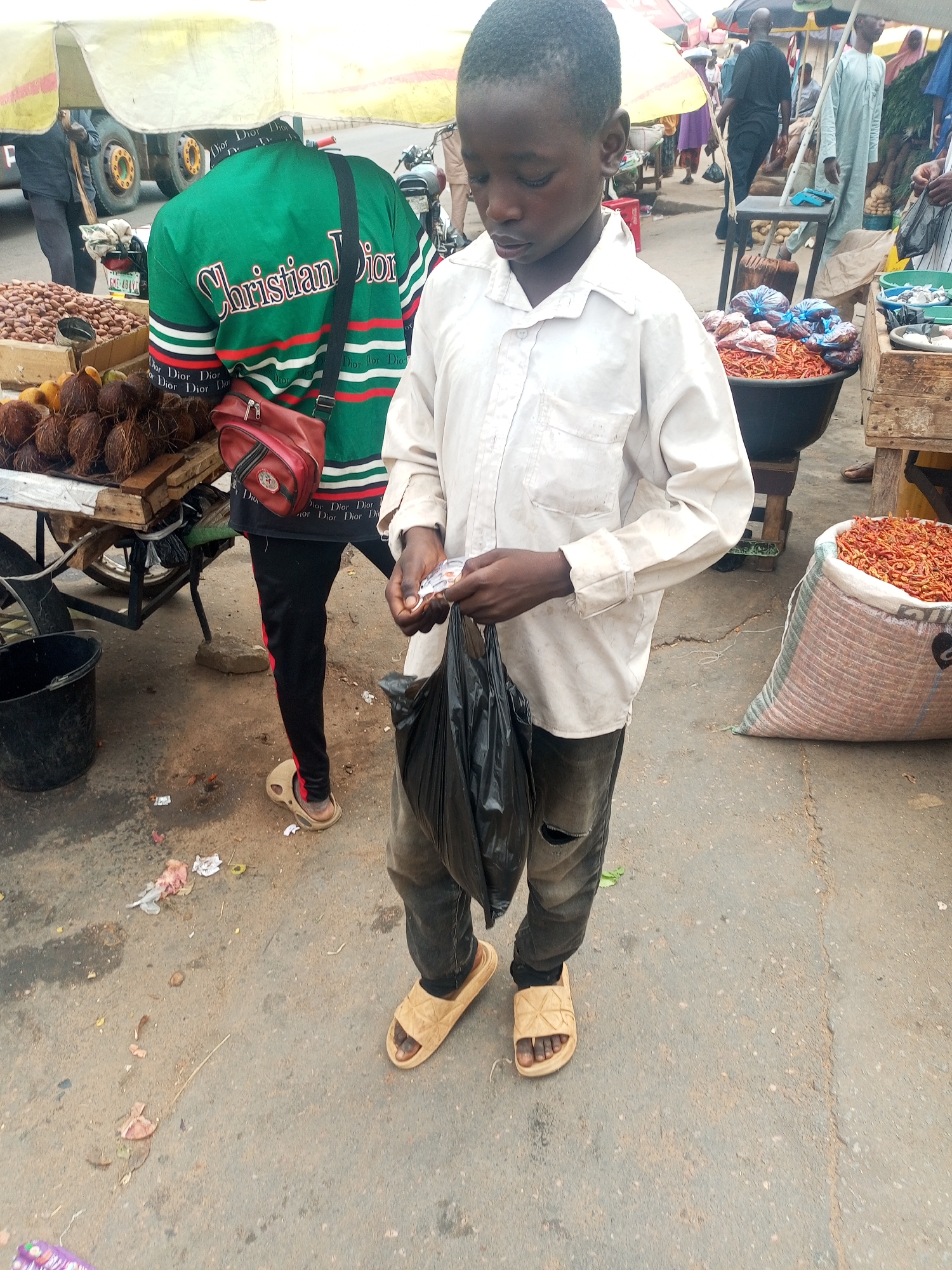 The scene depicts an outdoor market street in Unknown, Unknown. In the foreground, a young Black male, appearing to be an adolescent, stands looking down. He wears a long-sleeved white collared shirt, dark ripped jeans, and light tan slides. He holds a small white item, possibly money or a receipt, in his right hand and a black plastic bag in his left hand.
To his left and slightly behind, an adult Black male is seen from the back, wearing a green long-sleeved shirt with "Christian Dior" written in white script across the back, and "Dior Dior Dior" repeated on the lower back and sleeves. He carries a reddish-brown cross-body bag over his right shoulder and wears black track pants with red side stripes and beige Crocs-style clogs. He is positioned near a wooden market cart displaying piles of coconuts and a box of smaller brown nuts. This stall is shaded by a yellow and white striped canopy. A black plastic bucket rests on the ground near the cart.
To the right, a large black tub and a white sack are filled with dried red chilies, placed on the ground next to a wooden crate. The ground is an uneven, dirty paved surface with scattered debris. In the background, blurred figures of other people, some under market umbrellas, suggest a bustling environment. A truck and a motorcycle are partially visible in the far background. The scene is illuminated by bright natural daylight.