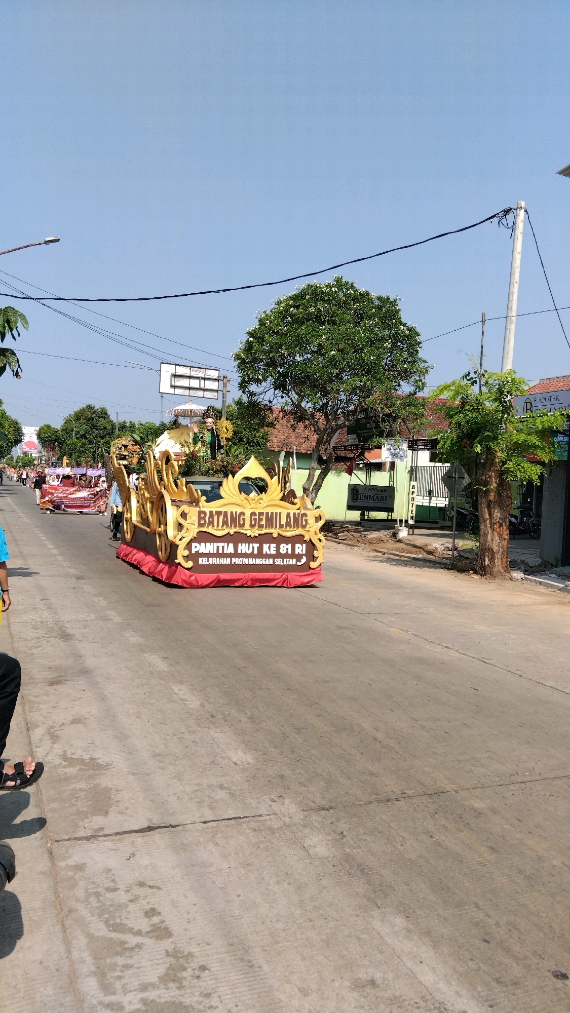 The image captures a street scene in Batang, Indonesia, likely during a parade or festival. A large, ornate float shaped like a golden chariot is the central focus. It is decorated with intricate carvings and topped with a golden crown. The float displays text that reads "BATANG GEMILANG" and "PANITIA HUT KE 81 RI KELURAHAN PROTONGGAN SELATAN."
The float is positioned on a paved road with visible tire marks and some wear. Several trees line the street, providing shade. In the background, buildings with signs are visible, including one that appears to be a pharmacy ("APOTEK"). The sky is clear and blue, indicating a sunny day.
The scene appears to be outdoors, on a street within a populated area. The presence of a float and the celebratory text suggest an event marking a significant anniversary, likely Indonesia's Independence Day (HUT RI). People are present on the float, though not clearly visible, and likely on the sidewalks or also participating in the procession. The overall atmosphere is festive and community-oriented.
