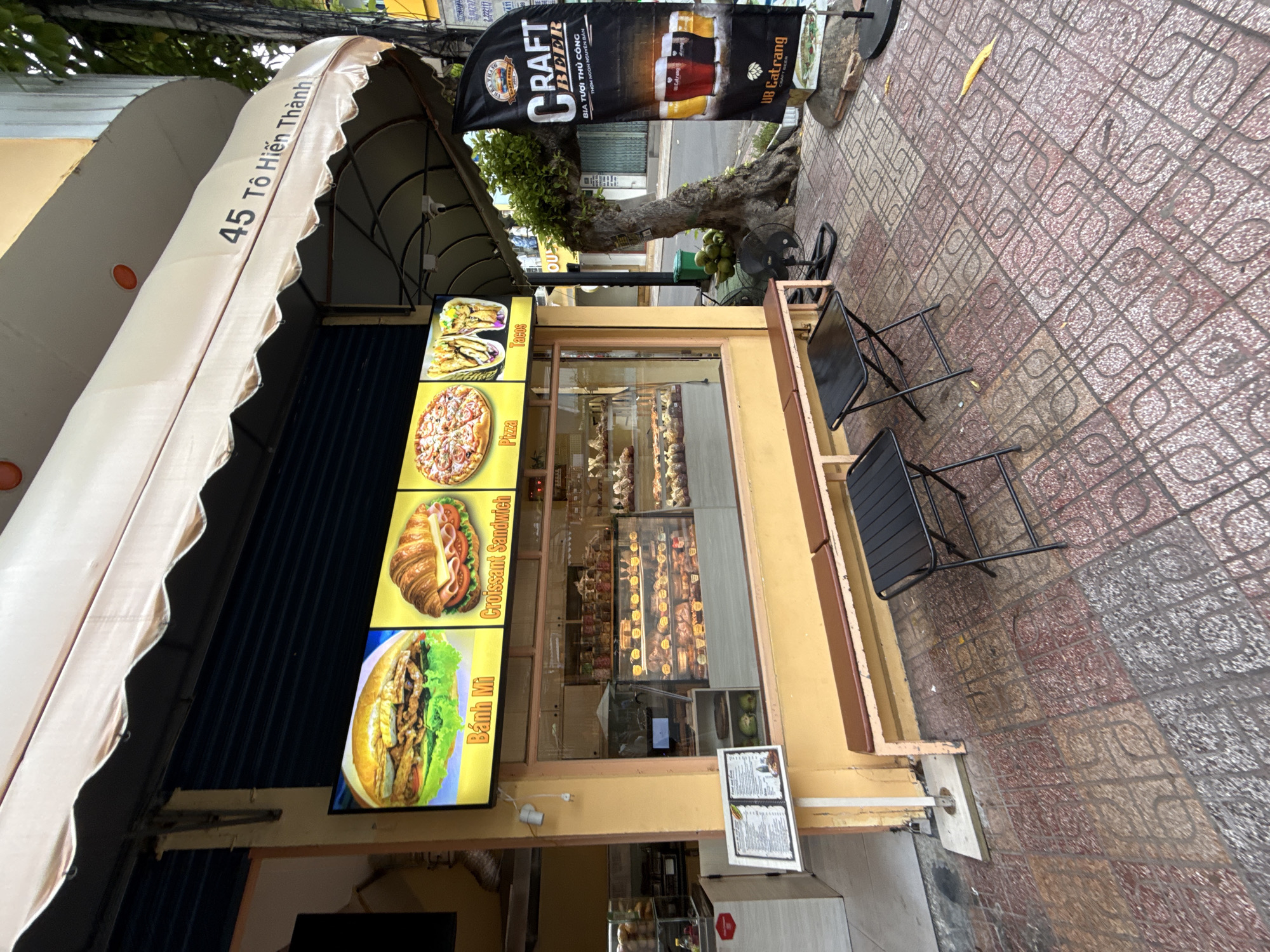 This image shows the exterior of a cafe located at 45 Tô Hiến Thành Street in Nha Trang, Vietnam. The cafe features a yellow facade with a prominent awning. A digital menu board above the storefront displays images of food items including Banh Mi, Croissant Sandwich, Pizza, and Tacos. Inside, a display case showcases various pastries and baked goods. Outside, a bench and two small black tables are set up on a tiled sidewalk, suggesting an outdoor seating area. A black flag banner with "CRAFT BEER" printed on it stands to the right of the entrance. The scene appears to be captured during the daytime on a cloudy day.