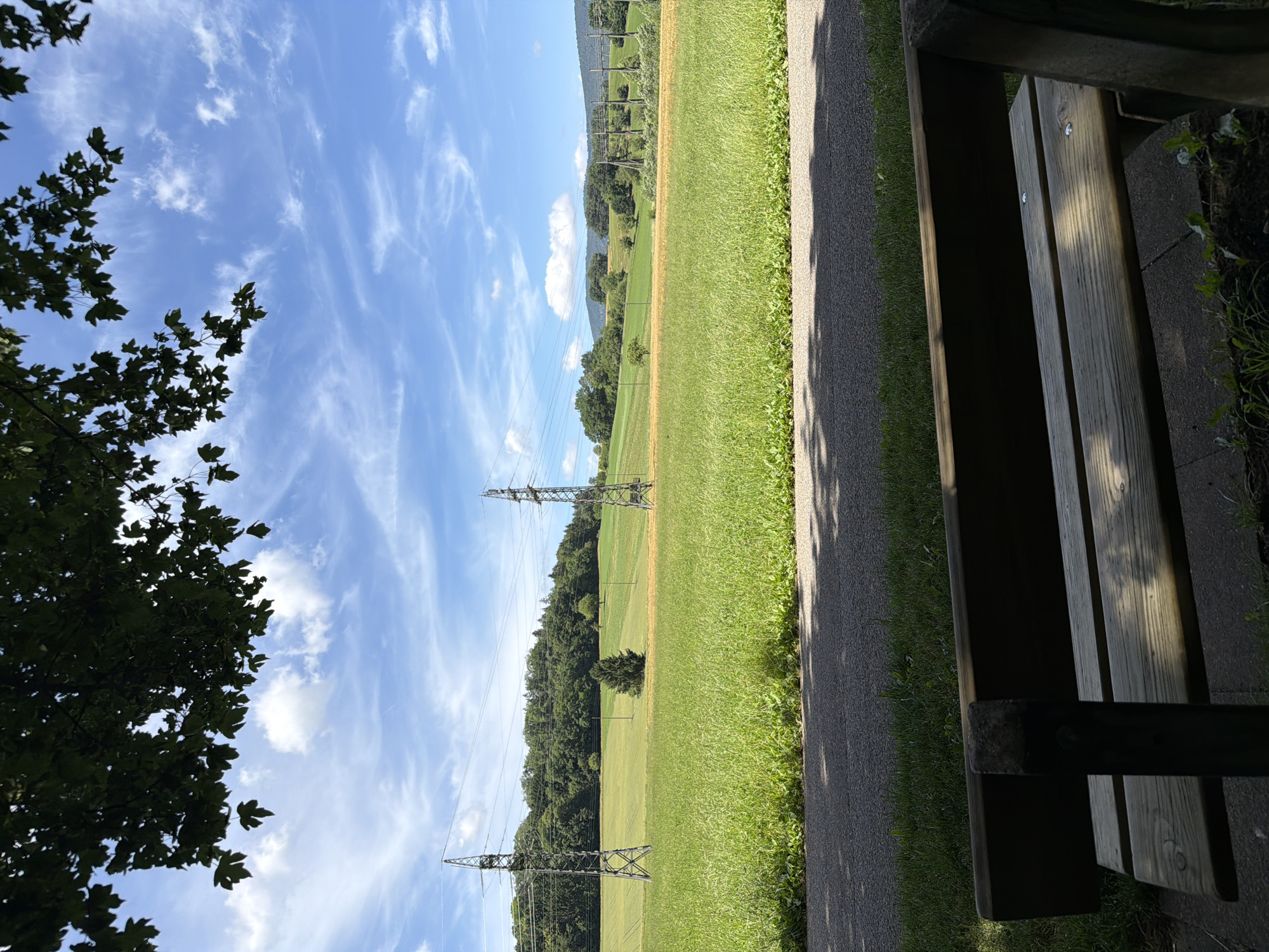 A park bench is in the foreground, with a paved path behind it. Beyond the path is a grassy field with two tall electrical towers. Further in the distance, a treeline forms a backdrop under a blue sky with scattered white clouds.