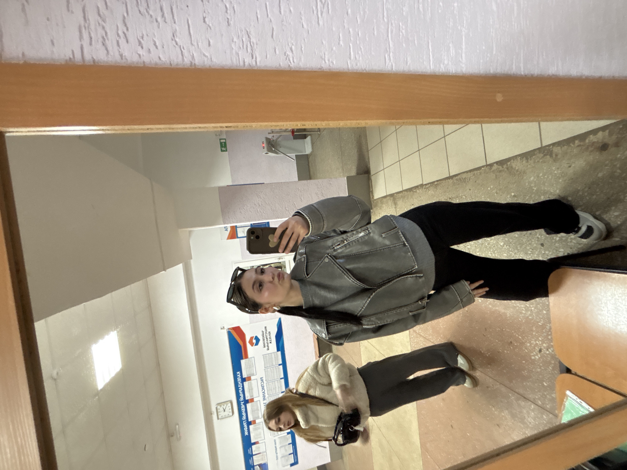 The image displays a mirror selfie taken by a young woman in what appears to be a public building, such as a school or administrative office, located in Chaikovsky, Russia. The primary subject, centered in the frame, holds a smartphone to capture her reflection, striking a playful pose with puckered lips. She is dressed in a grey faux leather jacket, a black top, black wide-leg pants, and light-colored sneakers with dark details. Sunglasses rest on her head.
Reflected to her left and further back is another young woman, looking down, wearing a cream-colored textured jacket, dark pants, and light shoes, and carrying a black handbag.
The background, visible through the mirror, reveals a hallway with white walls and a tiled floor. A prominent information board displays Cyrillic text, which includes Kazakh phrases such as "ЖОЛЫҢЫЗДЫ ТАҢДАҢЫЗДЫ" (Choose your path/way) and "КӨҢІЛІҢІЗ ЖАЗЫЛСЫН" (May your mood/spirit be written down). The word "БЕСІНШІ" (fifth) is also discernible. A wall clock is mounted near the information board. Further in the reflection, a green exit sign, a wall-mounted sink or water fountain, and a "WC" sign are visible.
The lighting indicates an indoor daytime setting. The scene conveys a casual atmosphere. A wooden frame surrounds the mirror, visible at the top and left edges of the image.