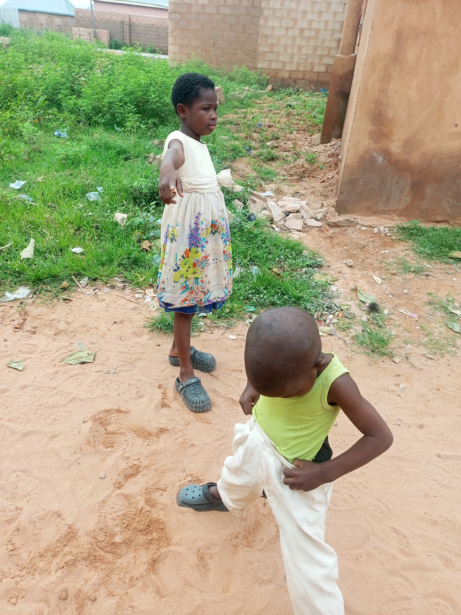 In the city of Tempure, Nigeria, two young children are observed in an outdoor setting characterized by sandy terrain and sparse vegetation. The child in the foreground, facing away from the camera, wears a light green tank top and cream-colored pants. This child's posture suggests engagement with something on the ground. Behind them, a second child, dressed in a light-colored dress adorned with floral patterns, stands facing the viewer and gesturing forward with their right hand. The background reveals elements of the urban environment, including brick structures and a tall, textured wall. The ground is a mix of sand and dry soil, with scattered debris such as leaves and small pieces of rubble.