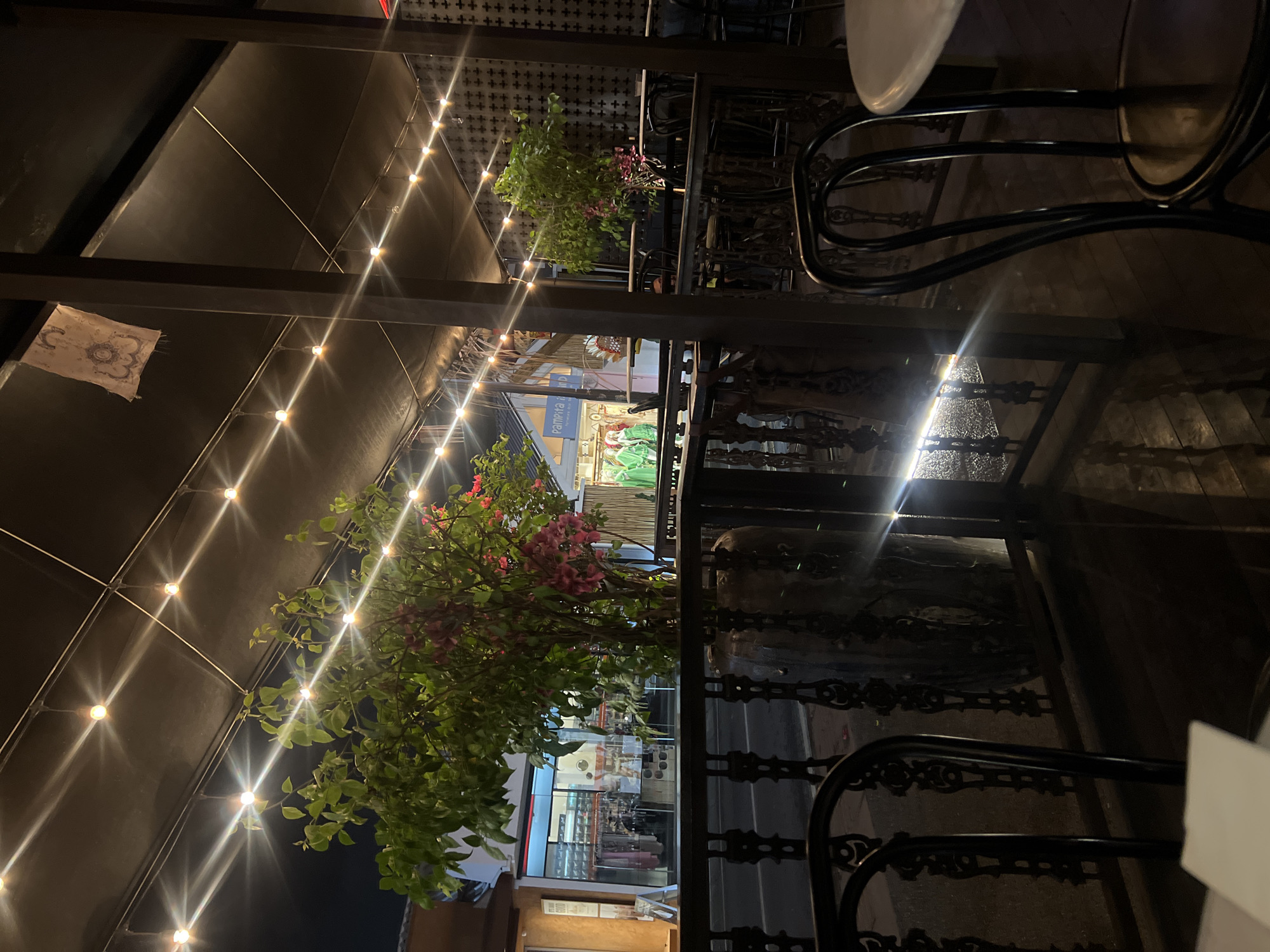 The image depicts an outdoor dining area at night, with string lights illuminating the scene. The setting appears to be a restaurant or cafe with an outdoor seating section, likely in North Kuta, Indonesia. The area is furnished with dark-colored metal tables and chairs, some of which are visible. A lush green plant with pink flowers is prominently featured in the foreground, adding a touch of natural beauty to the urban environment. The lighting from the string lights creates a warm and inviting ambiance. In the background, the facade of a building is visible, with some shops or businesses indicated by illuminated signs and displays. One sign reads "Pamora" in blue lettering. The overall impression is one of a pleasant evening setting, suitable for dining or socializing. The image is captured in a low-light environment, suggesting it was taken after sunset. The textures of the metalwork, foliage, and ceiling structure are visible, contributing to the depth and detail of the photograph.