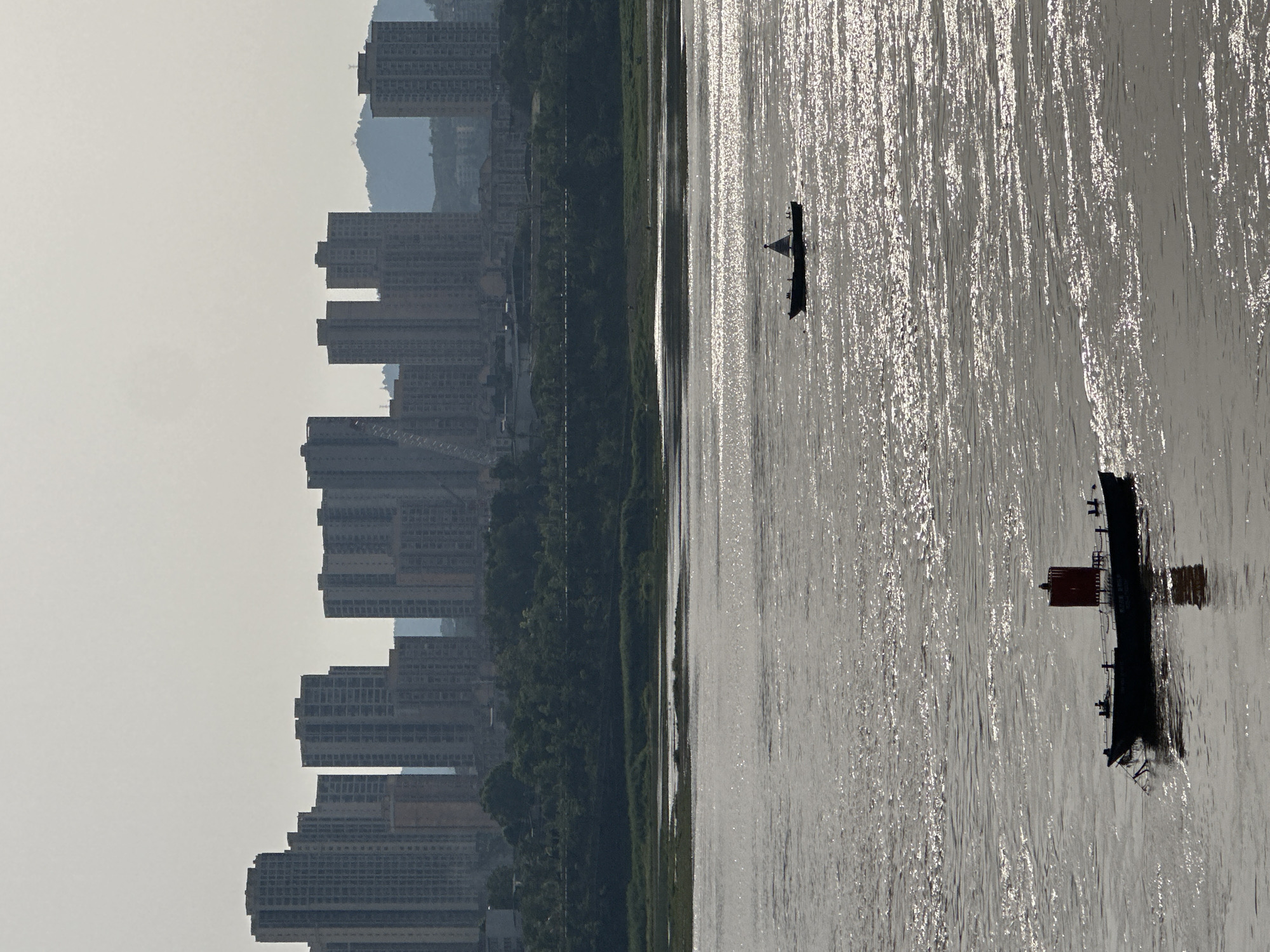 This image captures a wide panoramic view of a river and a city skyline under a hazy sky in Jiangjin, China.
The dominant feature is the wide river, which occupies the foreground and midground, appearing intensely bright and silvery due to direct sunlight reflecting off its surface. This creates numerous shimmering, vertical streaks of light across the water. Two boats are visible on the river. In the distance, a smaller boat is a dark silhouette against the glare. Closer to the viewer, a larger, darker vessel, possibly a barge or cargo boat, also appears silhouetted but with a distinctive reddish structure on its deck.
On the far bank, a continuous line of high-rise buildings forms a dense urban skyline. These buildings are uniformly dark grey and somewhat obscured by atmospheric haze, giving them a muted appearance. A strip of dark green trees and vegetation runs along the riverbank, separating the water from the buildings. Beyond the cityscape, faint, irregular outlines of hills or mountains blend into the pale, light grey sky, which itself appears hazy or overcast.
The bright reflections on the water indicate daytime, likely mid-morning to late afternoon, with the sun positioned to create significant glare. No text is visible in the image, and the scene conveys a sense of serene urban landscape.