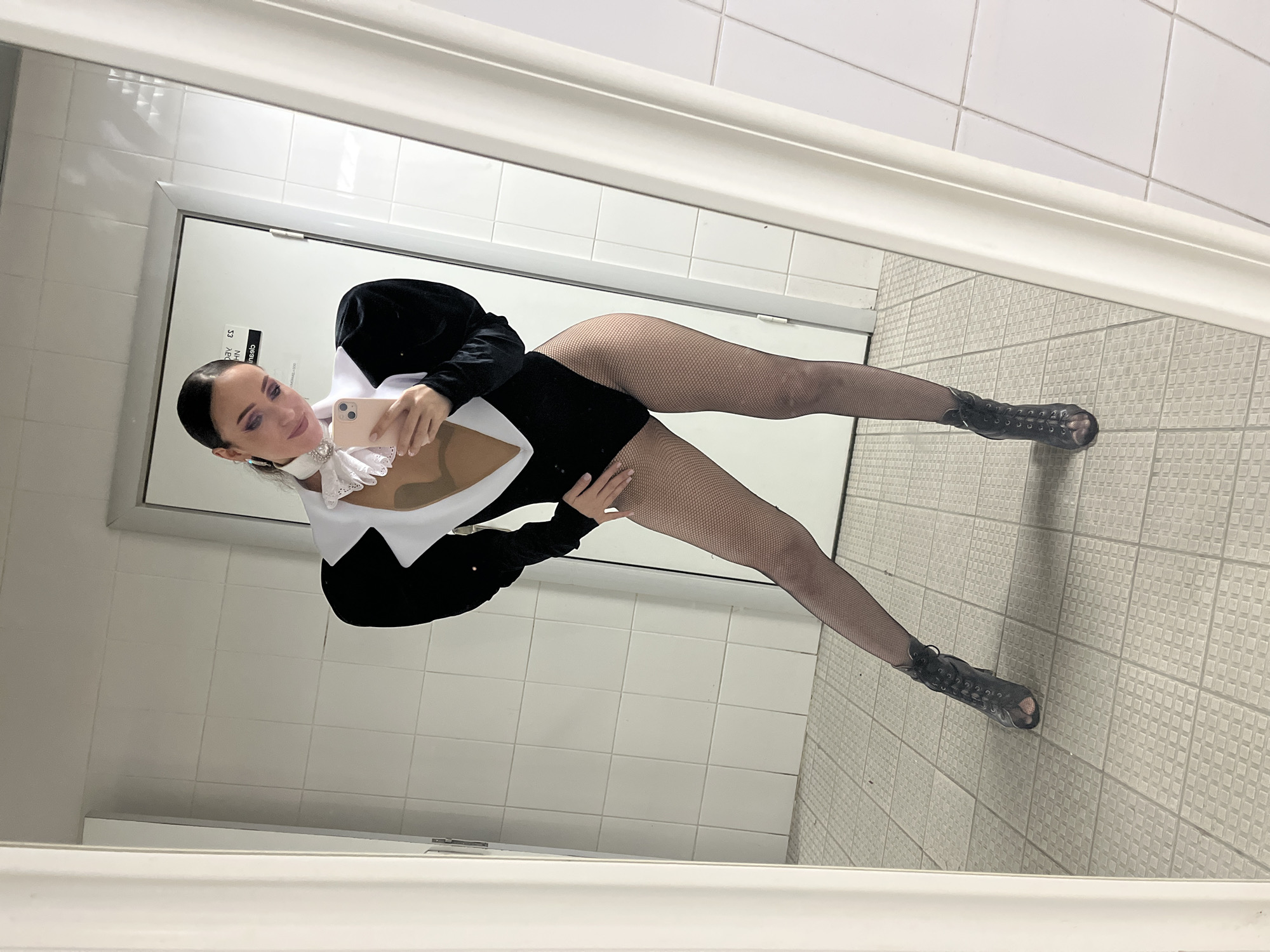 In a tiled restroom, a woman with dark hair pulled back poses for a mirror selfie. She wears a black, form-fitting leotard with puffed sleeves, a white plunging neckline, and a ruffled white collar adorned with a jewel. Her legs are clad in black fishnet stockings, and she sports black open-toe heeled boots. The room features white tiled walls and floor, and a white door is visible in the background.