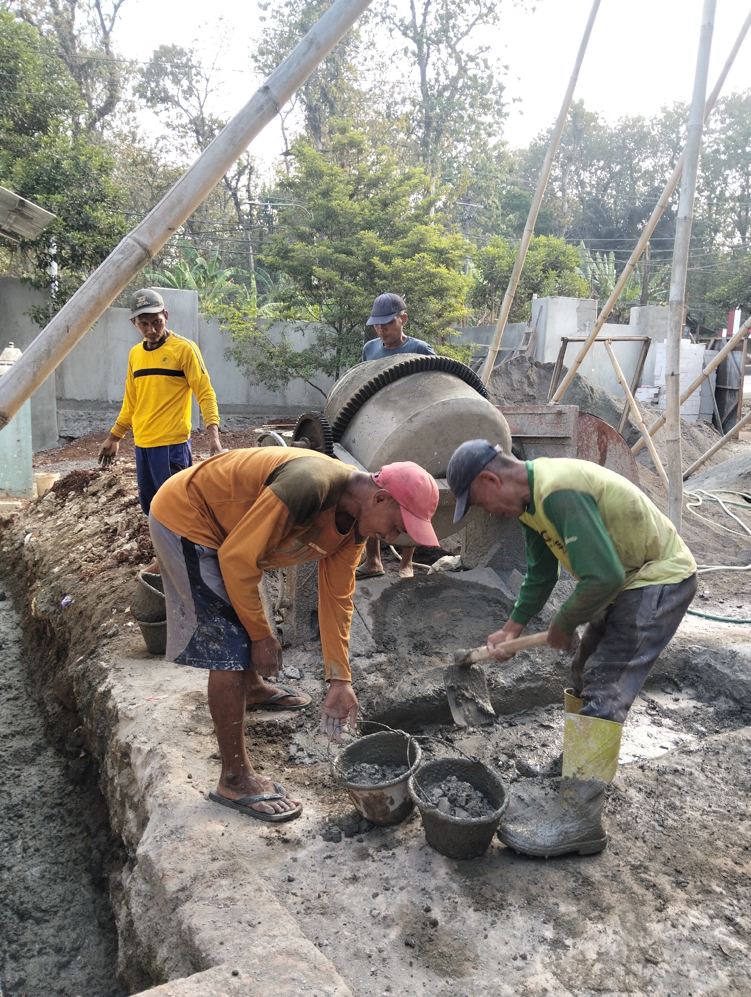 In Durenombo, Indonesia, four construction workers are engaged in mixing and pouring concrete. A large cement mixer is positioned in the background. In the foreground, two workers are bent over, one with an orange long-sleeved shirt and dark shorts, reaching into a bucket, and the other, wearing a yellow and green long-sleeved shirt and dark pants, is shoveling concrete. A third worker in a yellow long-sleeved shirt and blue pants stands nearby, observing. A fourth worker, wearing a blue shirt and dark cap, is partially visible behind the cement mixer. The ground is covered in wet concrete and construction debris, with several buckets scattered around. Tall bamboo poles and construction materials are also present in the environment.