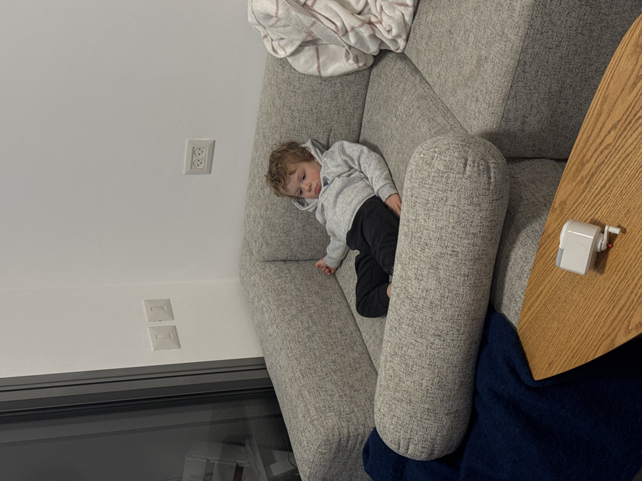 An indoor scene depicts a young child seated on a light grey modular sofa, likely in a residential living space in Tel-Aviv, Israel. The location is identified by the visible Type H electrical outlet on the white wall above the child. The child, a toddler with curly light brown hair, wears a light grey hooded sweatshirt with a dark emblem and dark trousers, appearing barefoot. The child is reclined with a somber or pouting expression, looking towards the right side of the frame. To the child's right, a white and pink striped blanket is crumpled on the sofa. A dark blue blanket is draped over the lower left portion of the sofa, beneath a cylindrical bolster cushion. On the far left, a large, dark-framed sliding glass door or window reveals a blurred reflection or exterior. Two white light switches are vertically aligned on the wall to the left of the sofa. In the foreground, a segment of a light brown, irregularly shaped wooden coffee table holds a small white mechanical device with a clear top and a red crank or handle.