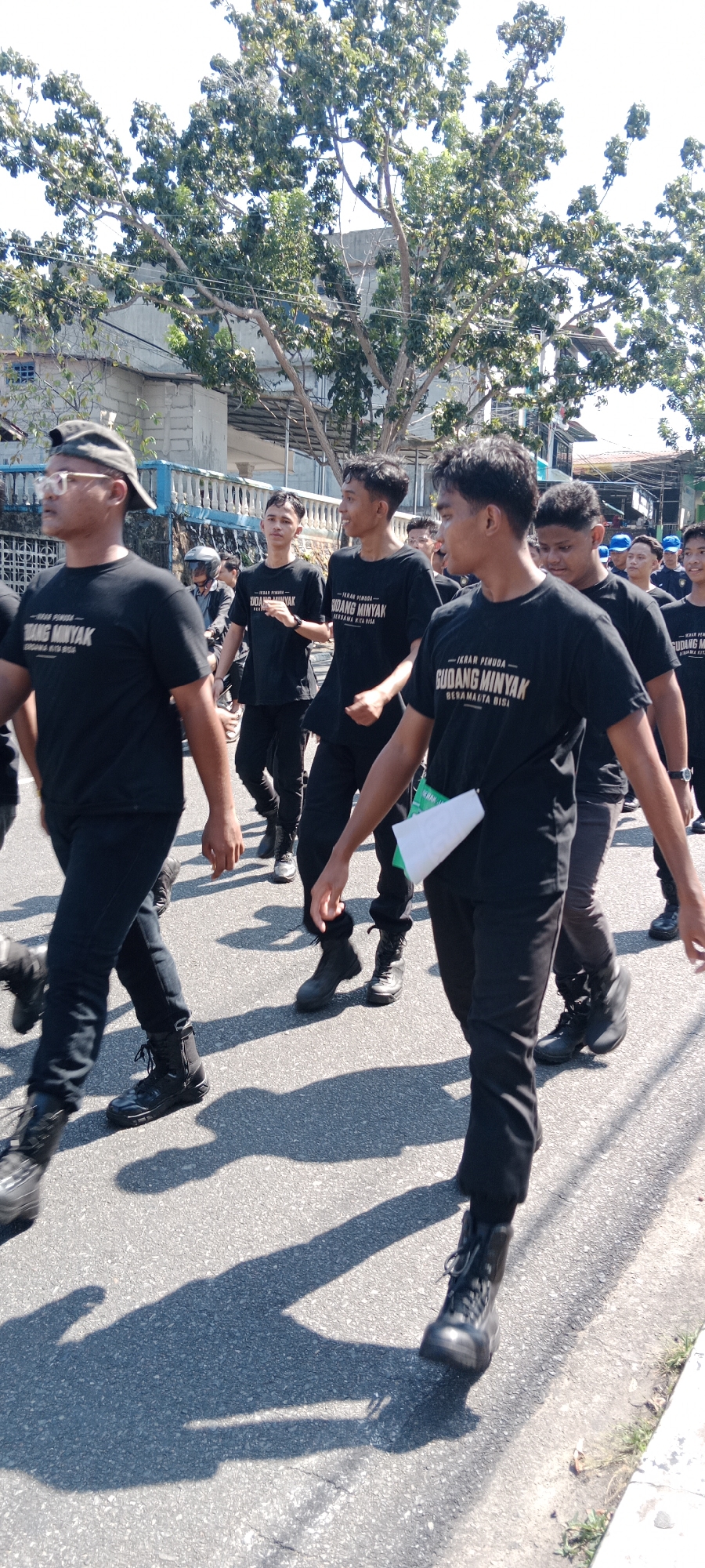 A group of young men in black t-shirts and black pants are walking on a paved street in Tanjung Pinang, Indonesia. The t-shirts have text on them that reads "IKRAR PEMUDA GUDANG MINYAK BERAMALITA BISA". The men are walking in the same direction and appear to be part of a procession or parade. Some of the men are wearing black boots, while others are wearing different types of footwear. In the background, there are trees, buildings, and a blue fence. The sun is shining, casting shadows on the street.