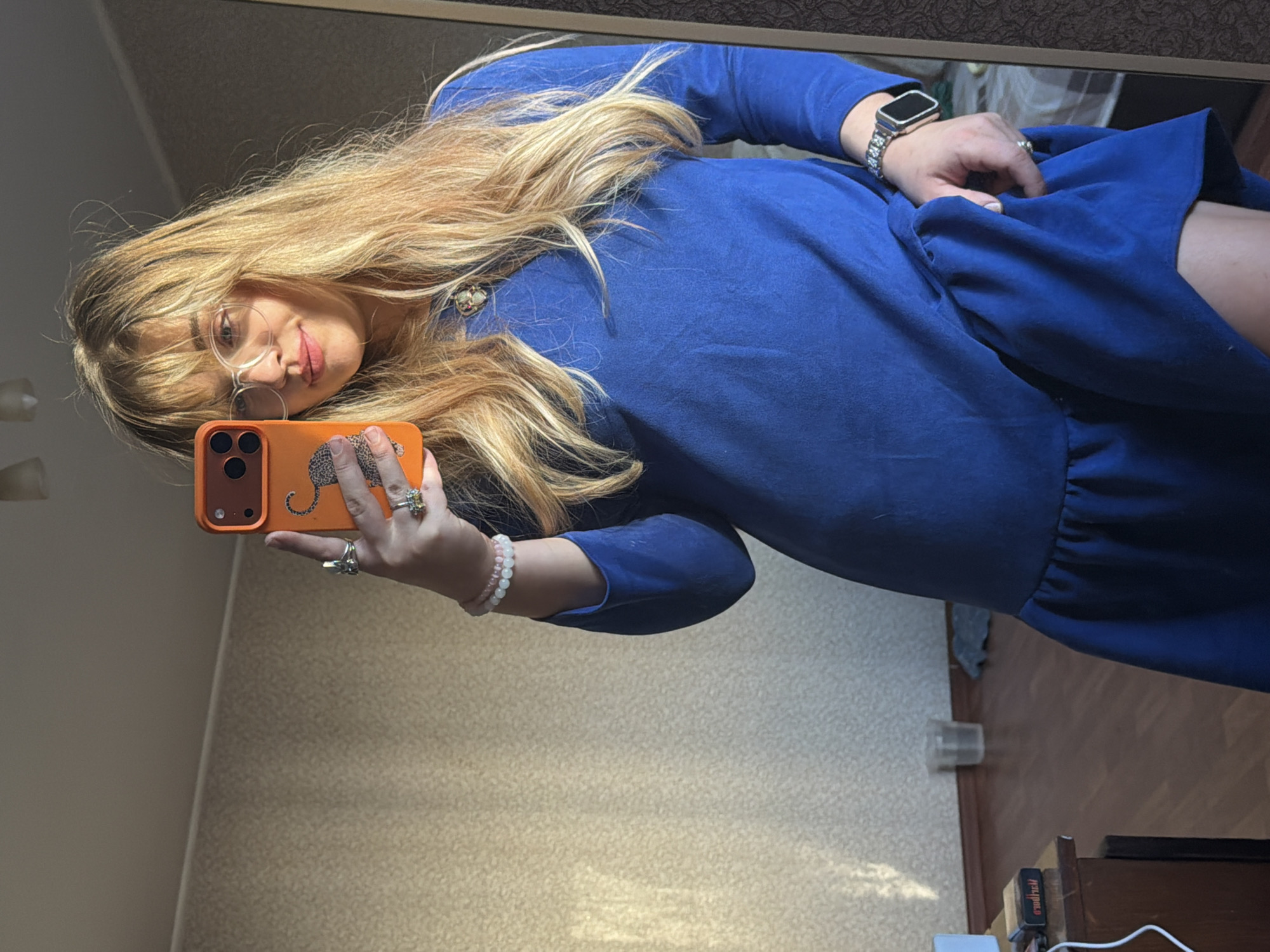 A woman with blonde hair is taking a selfie in a mirror. She is wearing glasses and a blue dress with long sleeves. She is holding an orange smartphone with a leopard print case. She is also wearing a bracelet and rings.
The background includes wallpaper and a wooden floor. It is not possible to definitively determine the location within the city of Naberezhnye Chelny, Russia from the given image alone.
