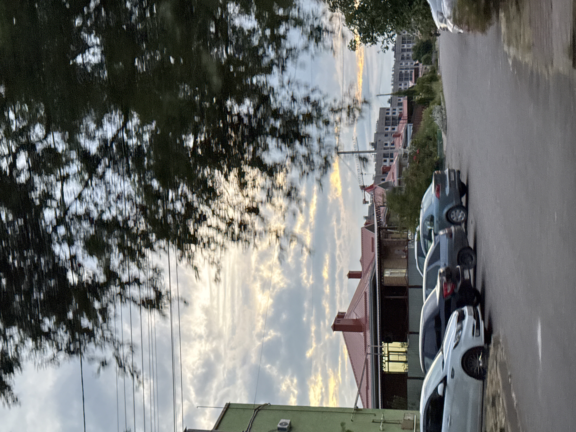 This media file depicts a street scene in Mikhaylovsk, Russia, likely during the late afternoon or early evening given the soft, warm light filtering through the clouds. The camera is oriented vertically, suggesting it was held in portrait mode.
The primary subjects are several parked cars lining a paved road. The cars are of various colors, including white, gray, and blue. The road appears somewhat wet, reflecting the sky and the surrounding environment. To the left of the road, lush green foliage from trees partially obscures the view, with their branches casting dappled shadows. Beyond the trees, a textured sky with elongated, wispy clouds is visible, some catching the golden hues of the setting sun.
In the background, there are residential buildings and construction sites. The buildings feature red tiled roofs and some appear to be multi-story apartment complexes. Construction cranes are also visible, indicating ongoing development in the area. One building has an air conditioning unit mounted on its exterior. The overall impression is of a suburban or semi-urban environment. There is no visible text in the media file.