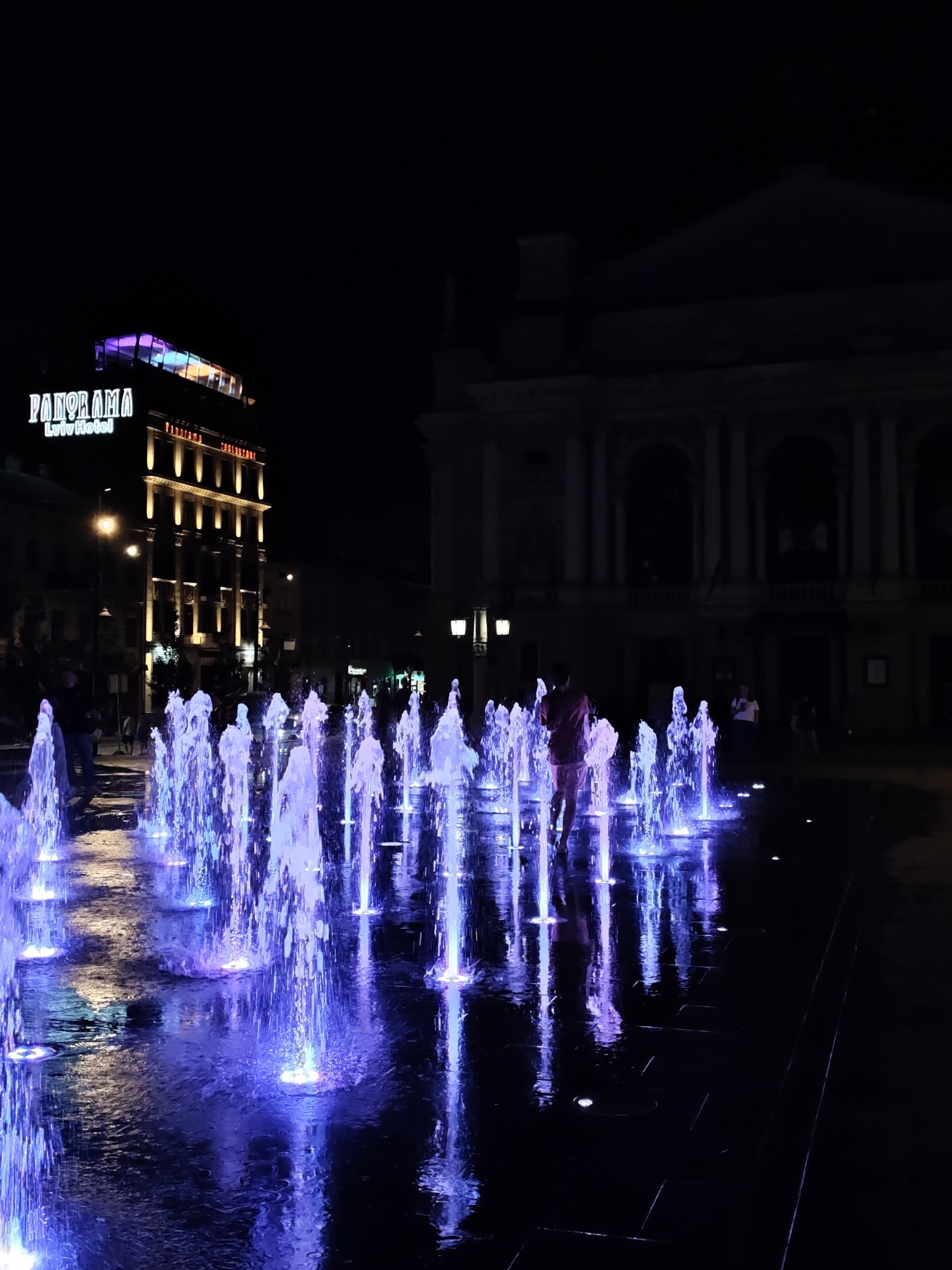 The image depicts a night scene in Lviv, Ukraine, featuring a vibrant water fountain display. The fountain's jets are illuminated with bright blue and purple lights, creating a striking visual effect on the wet pavement. In the background, a tall building with "PANORAMA Lviv Hotel" signage is visible, its facade lit up with yellow lights. To the right, the dark silhouette of a grand, arched building looms against the night sky. A few individuals are present, with one person, wearing a reddish-pink shirt, walking through the illuminated fountain. The scene is a lively urban public space at night, characterized by artificial light and the movement of water.