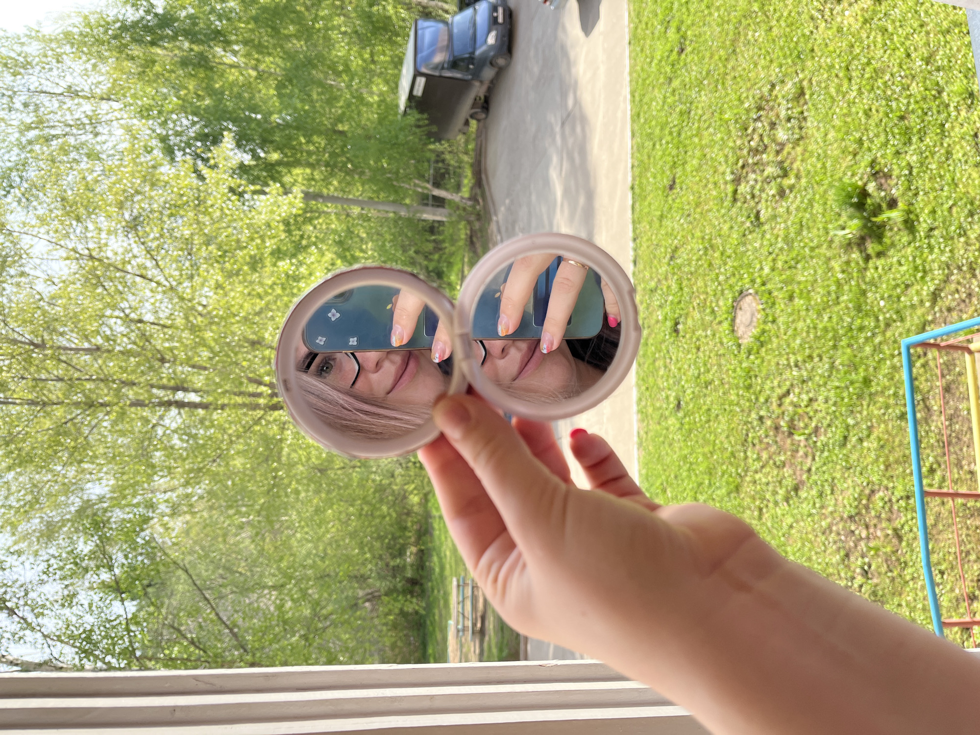 A person holds a double compact mirror near a window, reflecting their face. The person has glasses and light pink hair. The window looks out onto a scene with green trees and a grassy area. A vehicle is partially visible behind the trees. A playground structure is on the right side of the scene. The location is Chaikovsky, Russia.