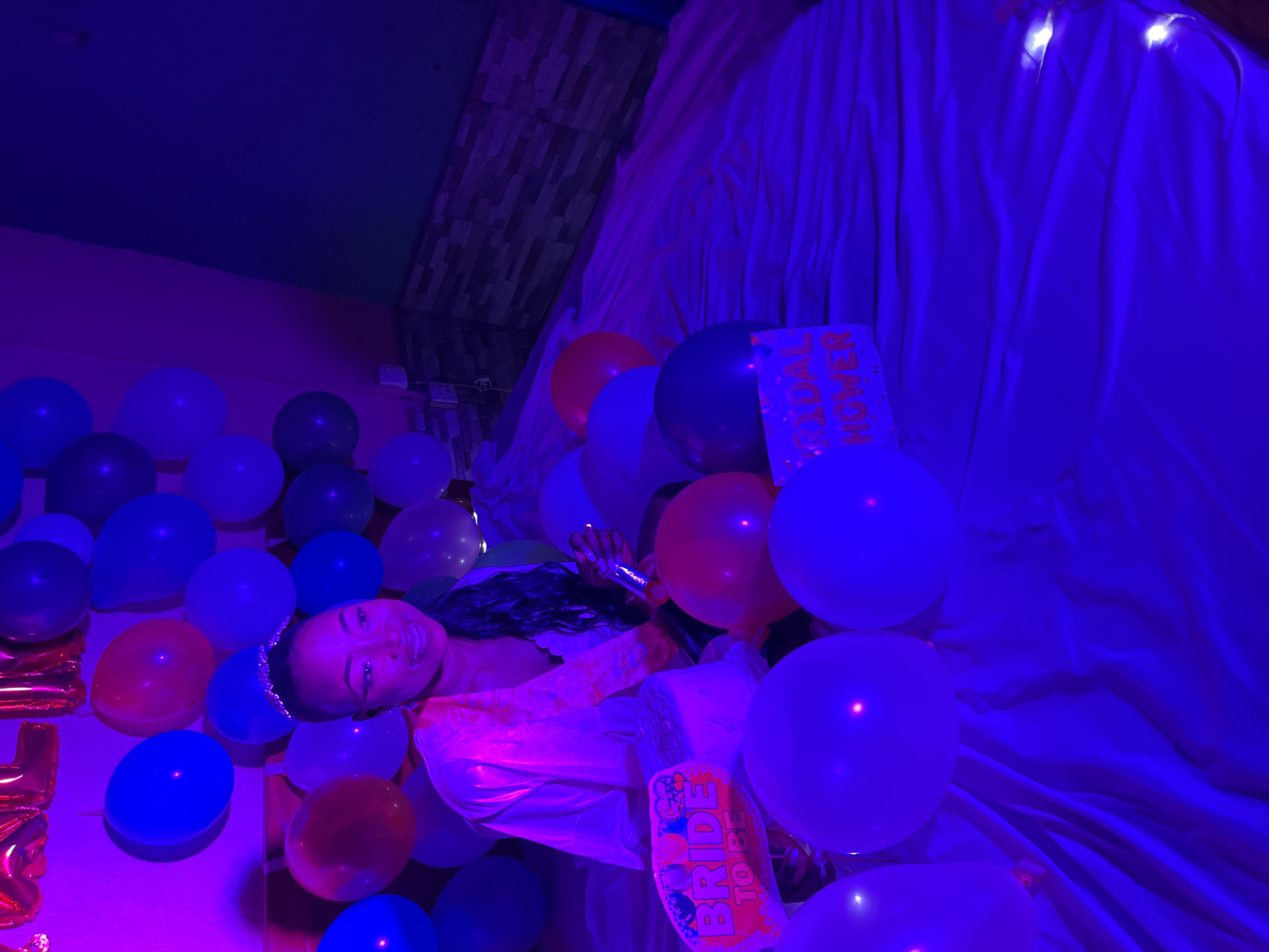 A woman lies among red, white, and blue balloons, holding a microphone and smiling in Akwanga, Nigeria. The backdrop is draped in white fabric, illuminated by blue lighting, with a brick wall and white lights visible at the top left. Two signs saying "Bridal Shower" and "Bride To Be" are present among the balloons.