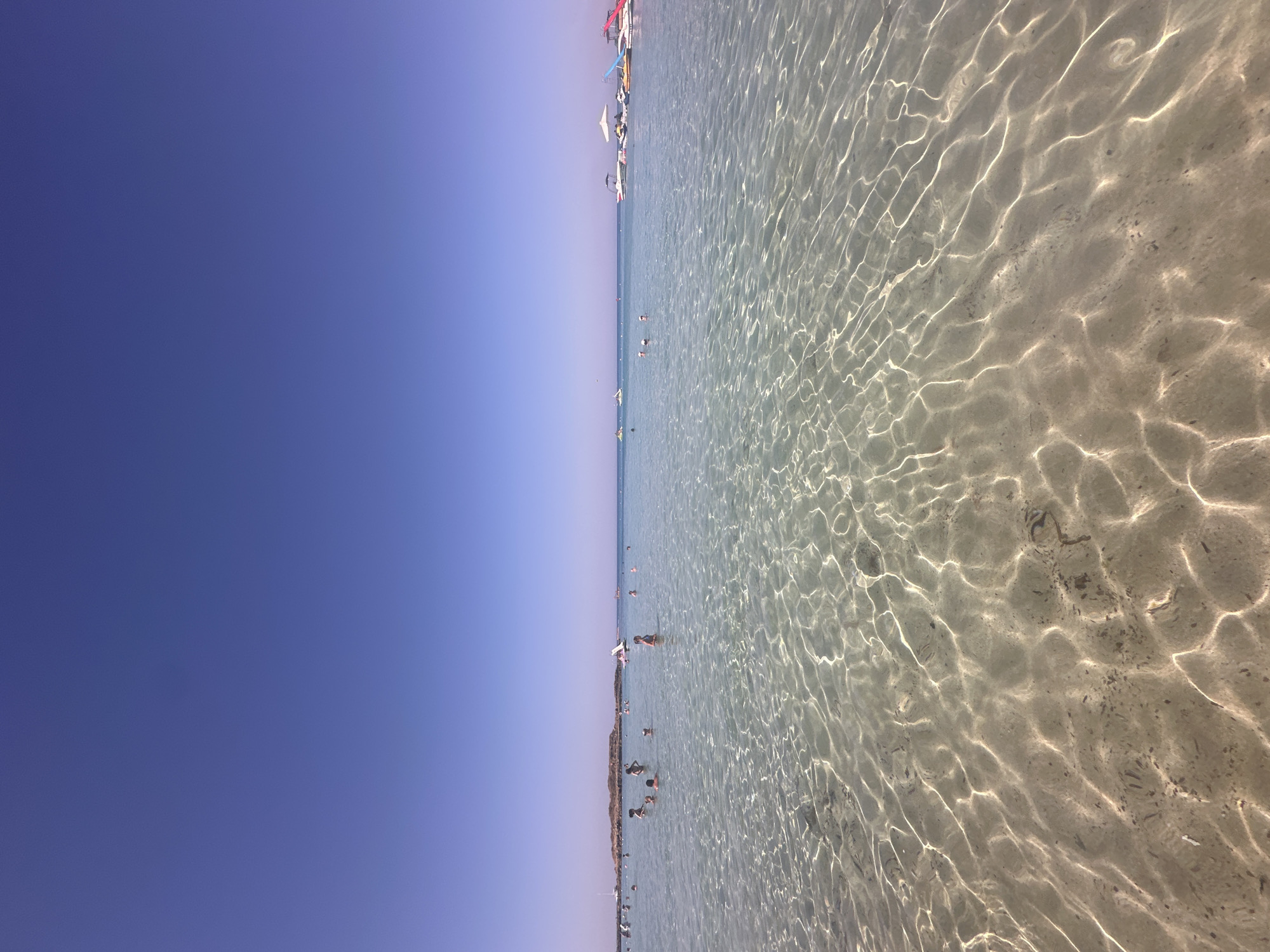 The image depicts a clear, shallow sea with a sandy bottom, under a bright blue sky. The water is calm, with sunlight creating shimmering patterns on the seabed. Several people are visible in the water, some swimming and others standing. In the background, there are boats docked near the shore and a distant island or landmass. The scene is consistent with a popular beach destination. This image is likely taken at Fig Tree Bay in Protaras, Cyprus, a well-known beach celebrated for its shallow, clear waters and golden sand.