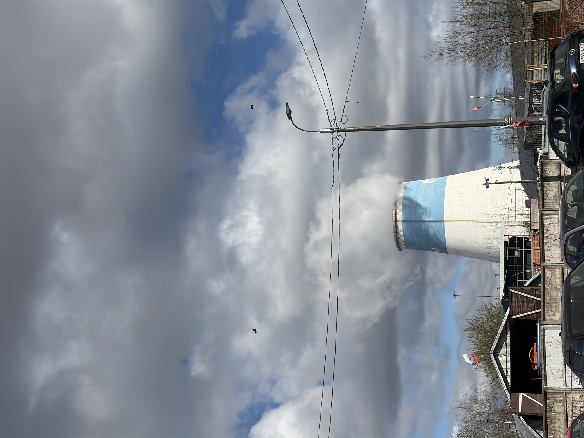 The image presents an industrial scene under a partly cloudy sky. A large, prominent cooling tower, primarily white with a broad blue band, dominates the left side of the frame, emitting a substantial plume of white smoke or steam into the atmosphere. The sky above is a mix of heavy grey clouds and patches of brighter blue.
Overhead, multiple power lines crisscross the frame, supported by a tall utility pole with a street light fixture, positioned slightly to the right of the cooling tower. Two dark birds are visible in flight against the cloudy backdrop.
On the right side of the image, several parked cars are partially visible, including a dark blue car in the foreground whose Russian license plate, "381YAA 178", is clearly discernible. The "178" region code corresponds to Saint Petersburg. Behind the vehicles and further to the right, there are various low-rise structures, likely garages or sheds, constructed from concrete and wood, with some graffiti visible on their surfaces. A Russian flag, with its white, blue, and red horizontal stripes, is flying on a pole amongst these buildings, confirming the location context. Bare tree branches are also scattered throughout the background.
The scene depicts an active industrial area, likely a power plant, during daytime hours, with functional infrastructure and elements of urban life.