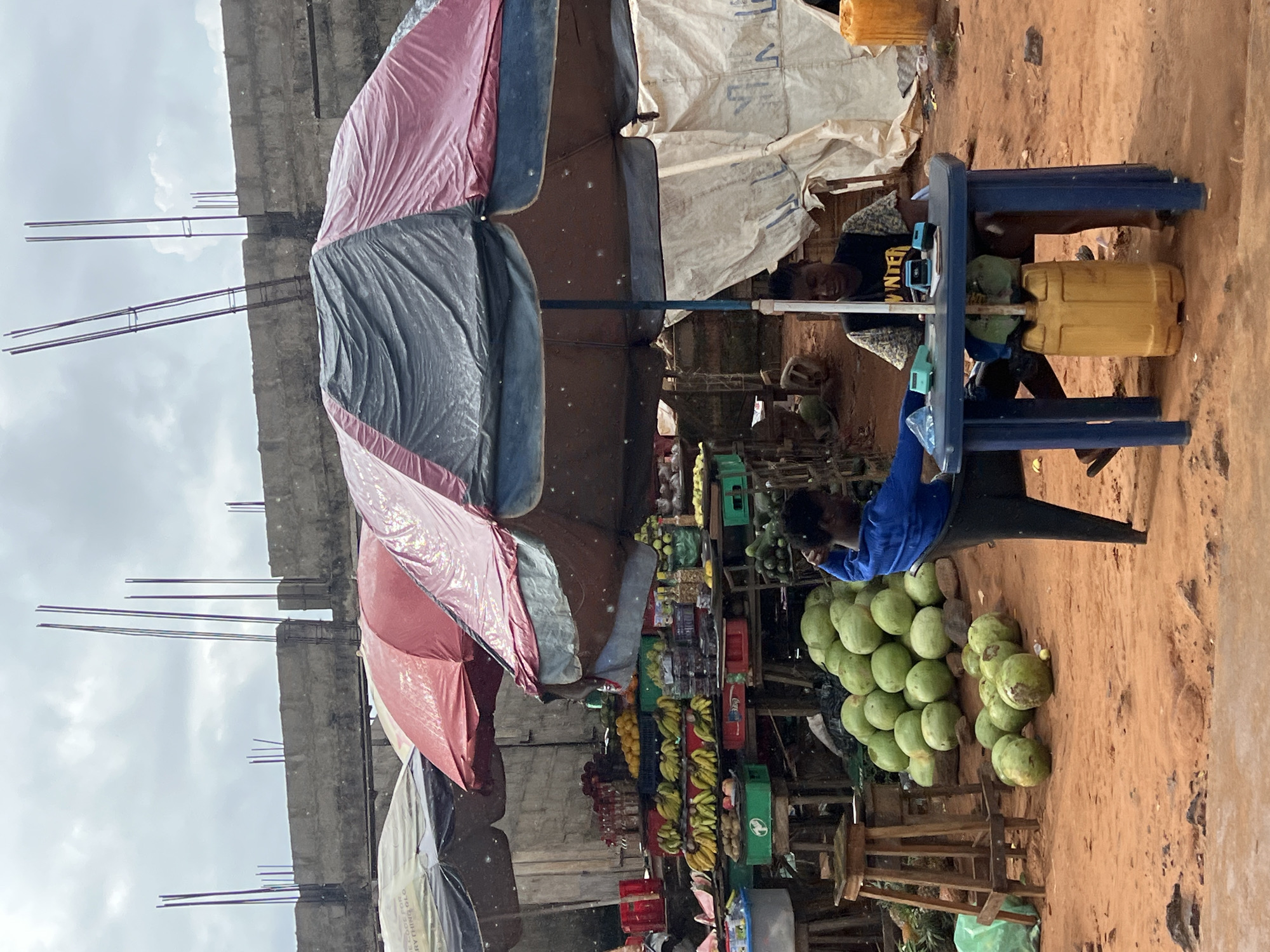 This is a medium shot of an outdoor market in Okpanam, Nigeria. Two young men are seated at a blue plastic table under a large, two-toned umbrella. The man on the left wears a blue long-sleeved shirt, and the man on the right wears a black t-shirt with "WINTER" printed on it. On the table are two small, blue digital devices, one open, and a blue plastic container. A large yellow plastic jerrycan is placed on the ground beneath the table. The ground is reddish-brown dirt.
Behind the table, a large pile of round, green fruits, possibly coconuts or watermelons, are stacked on the ground. To the left of the fruit pile, a wooden stool stands near a display of bananas and other fruits arranged on tables and in crates. Further to the left, a large umbrella shades another section of the market with more produce. In the background, a concrete wall with rebar protruding from the top suggests ongoing construction. The sky is overcast.