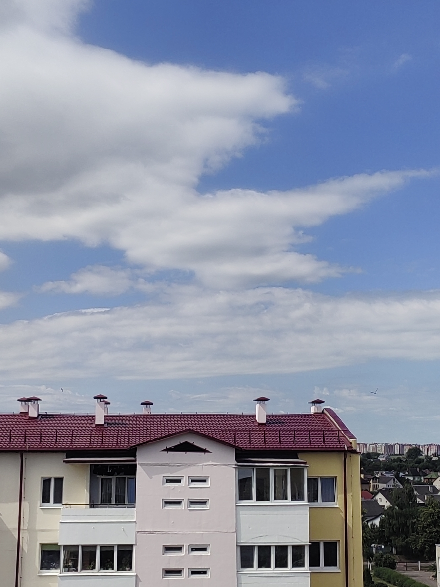 The image captures a view of a modern apartment building against a bright blue sky filled with fluffy white clouds. The building features a distinctive red-tiled roof with several chimneys. The facade of the building is painted in contrasting colors, with sections of yellow and light pink. It has multiple windows and balconies.
In the distance, a cityscape of smaller buildings and houses is visible, suggesting an urban or suburban setting. The overall impression is one of a clear, sunny day, possibly late morning or early afternoon, given the brightness of the sky and the clarity of the clouds. There are no people visible in the image, and the focus is on the architecture and the natural elements of the sky. A faint silhouette of a bird is visible in the sky, adding a touch of movement to the otherwise still scene. The location is not specified, but the architecture and the general appearance suggest a European setting.