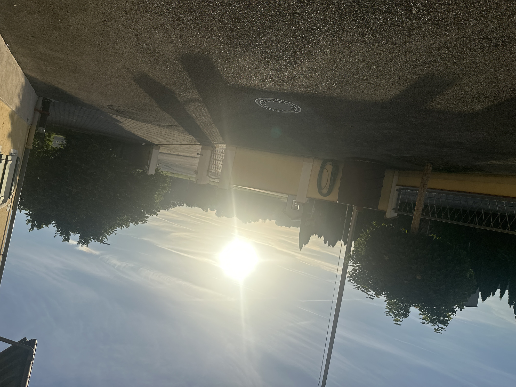 This is a wide-angle, eye-level, outdoor photograph taken on a sunny day in Mainburg, Germany. The image captures a residential street scene with a focus on a driveway leading to a building. In the foreground, a paved asphalt driveway occupies the majority of the frame, with the sun casting long shadows across it from the left. A circular manhole cover is visible in the driveway, slightly off-center to the left.
To the left of the driveway, there is a low, yellow-painted wall topped with a metal fence. A young tree with dense green foliage stands just behind this wall. Further back, a row of dark green, conical trees is visible. A tall, slender pole, possibly a flagpole or utility pole, extends vertically along the left edge of the image, partially obscuring the bright sun.
The sun is prominently positioned in the upper center of the frame, appearing as a brilliant, almost blinding white orb. Its rays create lens flare effects. Wispy white clouds are scattered across the bright blue sky.
In the mid-ground, a yellow building with white trim and a dark roof is partially visible on the right side of the image. Adjacent to the building is a paved walkway or driveway area. A metal gate, partially open, leads further into the property. Green foliage, likely trees and bushes, forms a backdrop on the right side of the image. A coiled green hose is attached to the yellow wall on the left side of the property. The scene depicts a quiet, residential area bathed in the warm light of the sun.