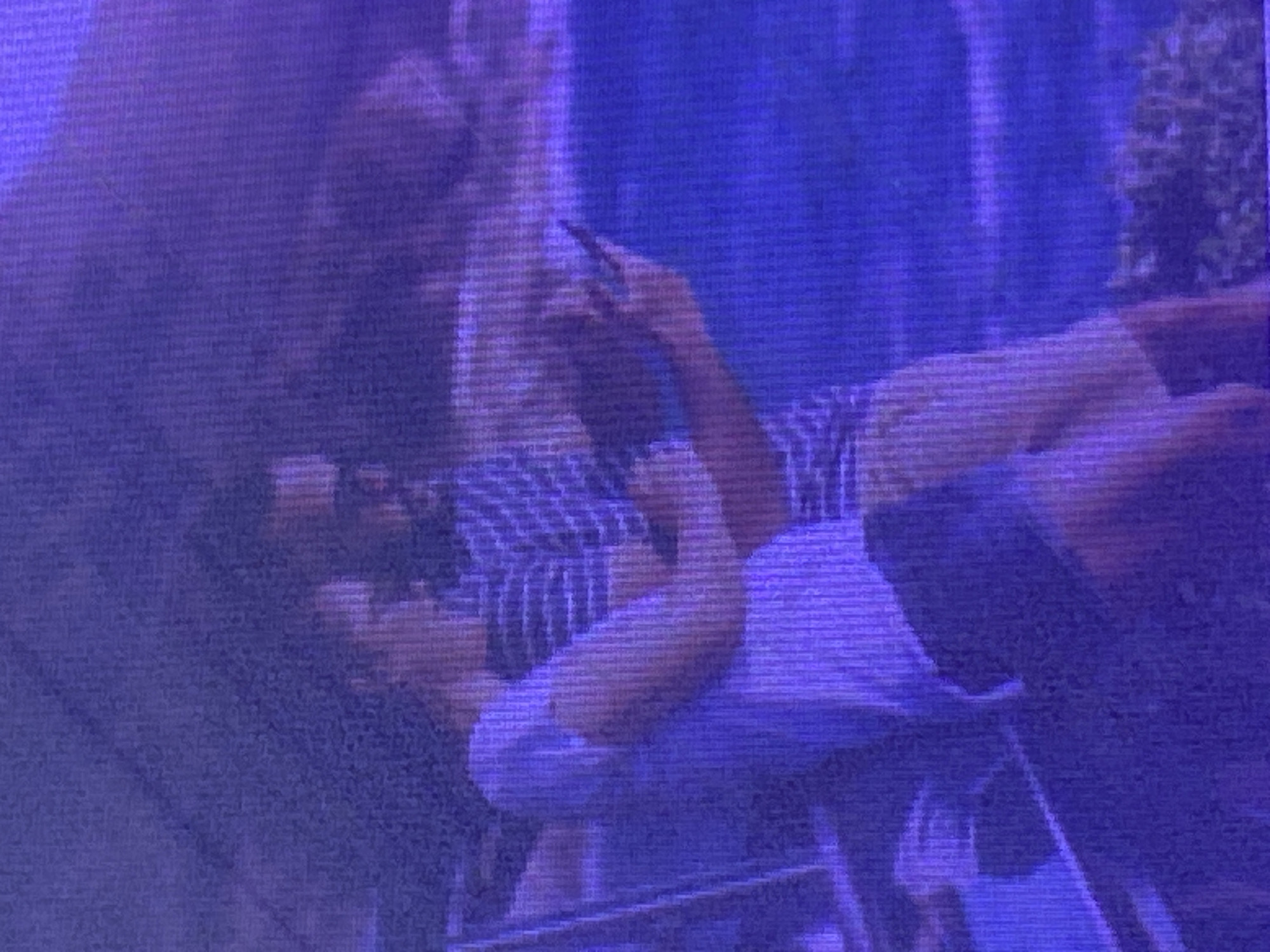 The image shows two people in a close-up shot, with the overall hue of the image being a deep purple and blue, suggesting it might have been taken at night or with a specific lighting filter. One person is lying down or reclined, wearing a blue and white striped shirt and what appears to be shorts. Their head is resting on a surface, and they are holding a mobile phone, seemingly engaged with it. The other person is positioned behind and partially above the first, their head bent down as if in conversation or interaction.
The scene appears to be outdoors, possibly in a garden or a park, with blurred greenery visible in the background, hinting at a natural setting. The quality of the image is grainy, making it difficult to discern fine details about the individuals' faces or the surrounding environment. However, the posture and proximity of the two individuals suggest a relaxed and intimate interaction. The presence of the mobile phone indicates a contemporary setting.
Given the location context of Ikorodu, Nigeria, this could be a personal moment captured during an evening gathering or leisure time. The dim lighting and the focus on the individuals create a sense of privacy. There is no visible text in the image.