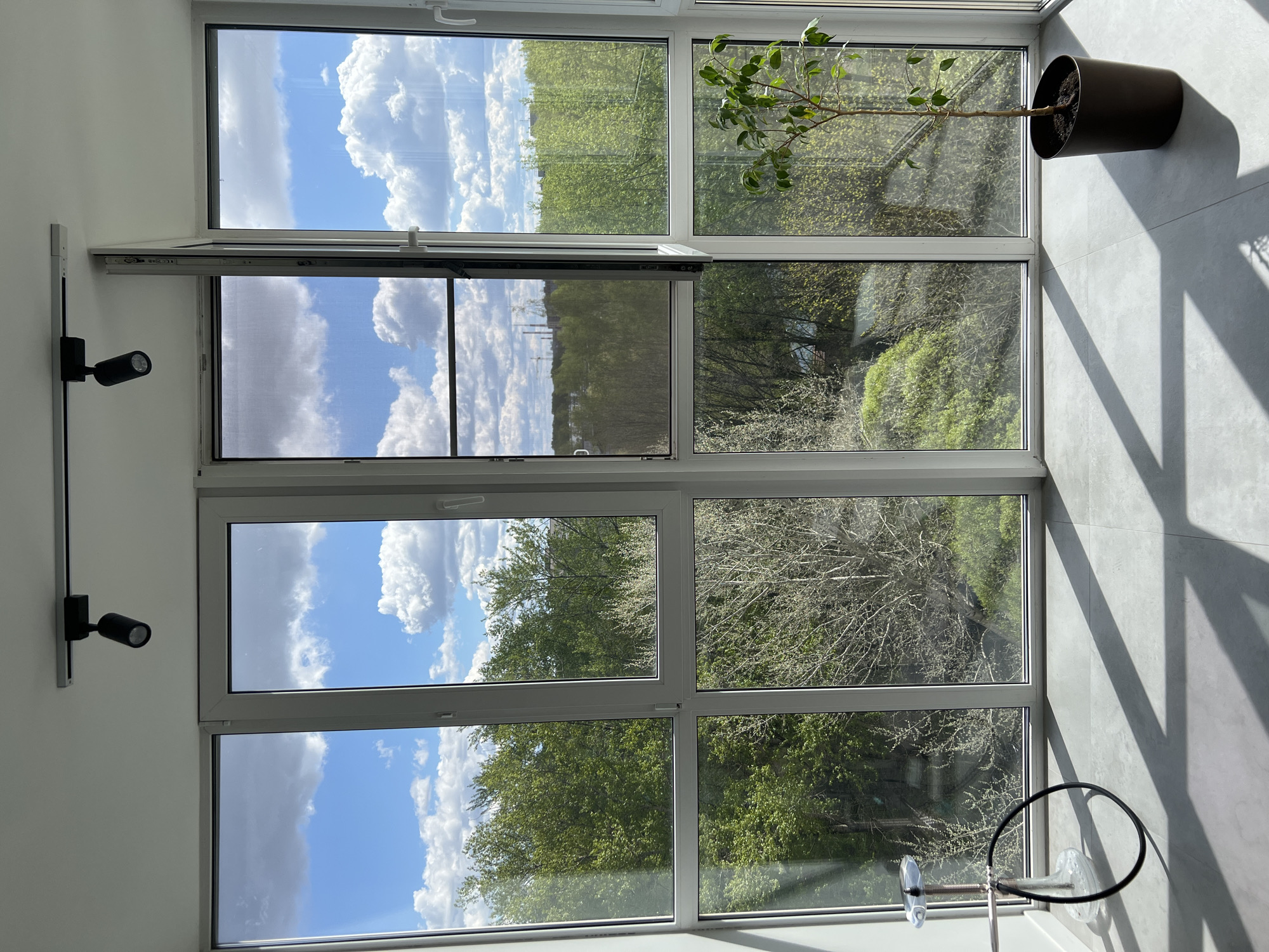The image shows an interior view of a room with a large window offering a view of a cityscape, possibly Vologda, Russia. The window is divided into multiple panes and reflects a partly cloudy sky. Outside, trees are visible, with green foliage suggesting springtime or early summer. A potted plant sits on the floor near the window, casting a shadow on the grey tiled floor and wall. A track lighting system with two black spotlight fixtures is mounted on the wall to the left. A section of the window is open with a screen. The scene appears to be indoors during daylight hours. A black circle attached to clear glass is at the bottom of the view.
