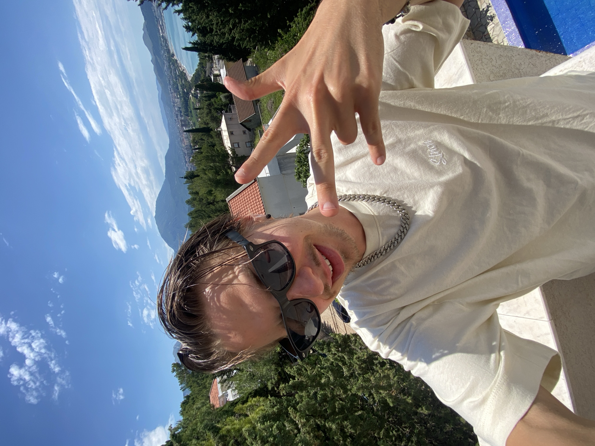 A man is posing for a selfie in Šušanj, Montenegro. He is wearing sunglasses and a white t-shirt, and a silver chain around his neck. His hand is raised in a gesture. Behind him is a view of the city, mountains, and the sea. The sky is blue with scattered clouds. Trees are visible in the foreground. Part of a blue tiled structure is in the upper right corner.