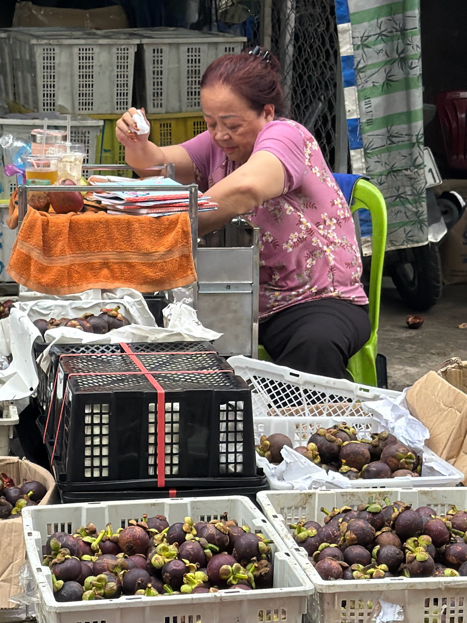 A middle-aged Vietnamese woman with dark hair pulled back is seated on a bright green plastic chair at a market stall in Nha Trang, Vietnam. She is wearing a pink t-shirt with a floral pattern and black pants. Her attention is focused on a small white object in her right hand, which she is holding near her face. In front of her is a metal cart with various items, including stacks of colorful magazines or books, a glass of amber liquid, and an orange towel draped over the front.
Surrounding the stall are numerous white and black plastic crates filled with mangosteen fruit. Some crates are stacked, and others are placed on the ground. The background reveals a chaotic, outdoor market environment with other stalls, a motorcycle, and a chain-link fence. The overall impression is of a typical busy market scene in Vietnam.