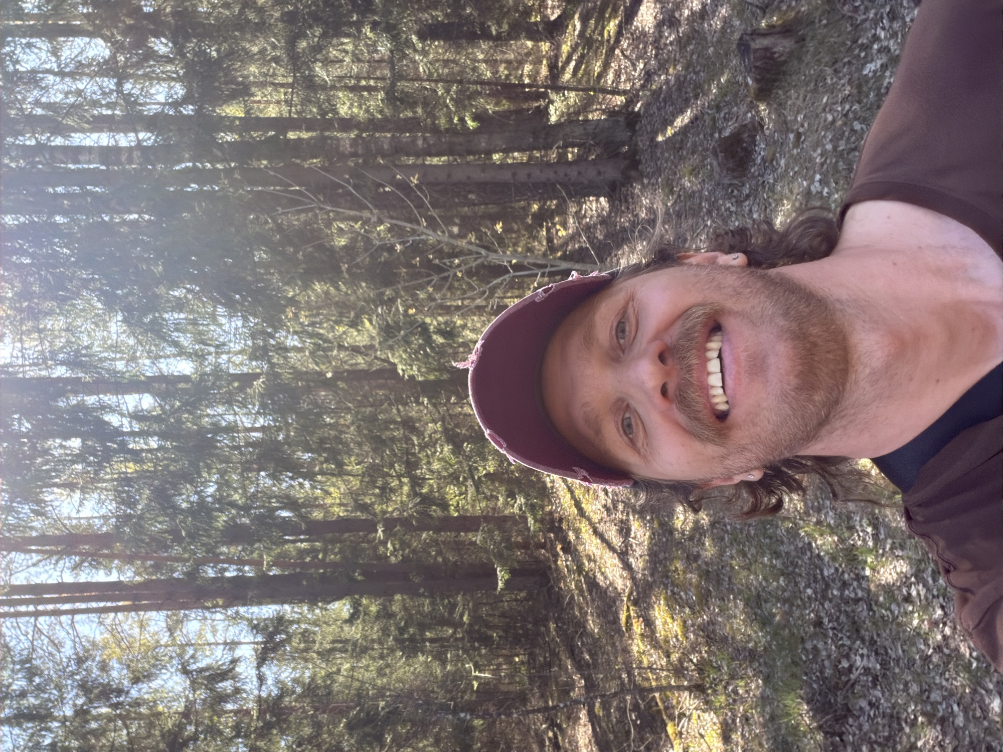 A man with a beard and a maroon cap is smiling. The background consists of many thin trees in a forest environment. The ground is covered with leaves and vegetation. The scene appears to be in a wooded area of Камянюцкі сельскі Савет, Belarus.