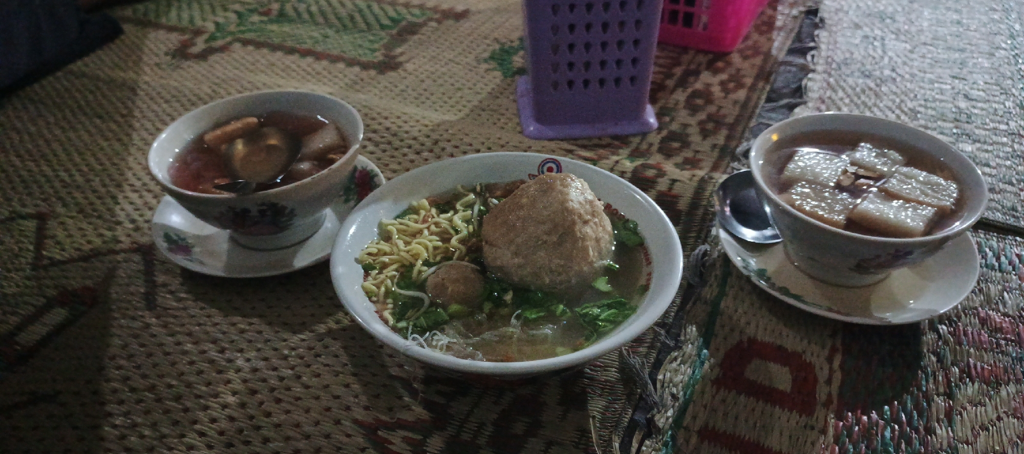 In the city of Sidoagung, Indonesia, a close-up, overhead view captures a dining scene set on a woven mat. Three bowls of food are present. The bowl on the left contains a broth with what appears to be meatballs or fish balls and possibly fried wonton pieces. To its right, a larger bowl holds a noodle dish featuring a large, round meatball, smaller meatballs, noodles, and green vegetables. The bowl on the far right contains a light-colored broth with several rectangular pieces of tofu or starch, and some almond slivers. A spoon rests in the bowl on the right, and another spoon is partially visible in the bowl on the left. A purple plastic container is situated in the upper right quadrant of the image. The lighting suggests an indoor setting, possibly a casual restaurant or home environment.