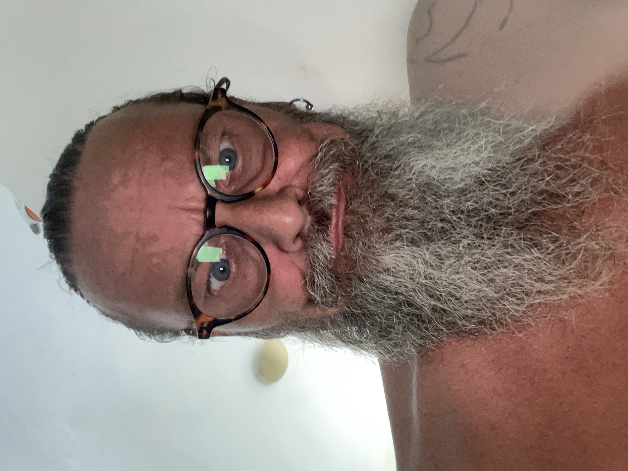 The image is a close-up, upside-down selfie of a man with a full, graying beard and long, dark hair pulled back. He is wearing round, tortoiseshell-patterned glasses. His eyes are blue and appear to be looking directly at the camera. The man has reddish-brown skin, and the lighting suggests it might be daytime, possibly indoors, given the diffused light and the presence of a light-colored, circular object on a white wall in the background, which could be a light fixture or a decorative element. The overall setting appears to be a bathroom or a similar indoor space with white walls. There is a faint tattoo visible on the man's upper arm or shoulder on the right side of the frame. The man's expression is neutral. No text is visible in the image. The location context is Netanya, Israel.